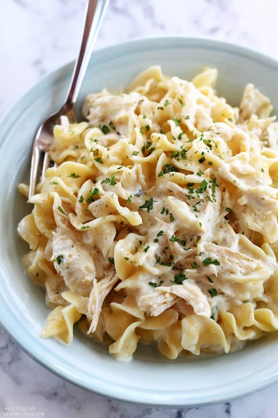 A close-up of creamy chicken noodle dish served in a white bowl placed on a white marbled surface. The dish has three main layers: at the bottom is a thick white cream sauce, on top of it a layer of soft cooked egg noodles with a slightly wavy texture and pale yellow color, and scattered throughout and on top are pieces of shredded chicken mixed well with the noodles. The whole dish is sprinkled with small green herb flakes for garnish. A fork is resting inside the bowl, ready to eat. photo taken with an iphone --ar 2:3 --v 7 - Crockpot Chicken and Noodles, easy slow cooker chicken dinner, Creamy chicken noodle casserole, make-ahead comfort food, family-friendly crockpot meal