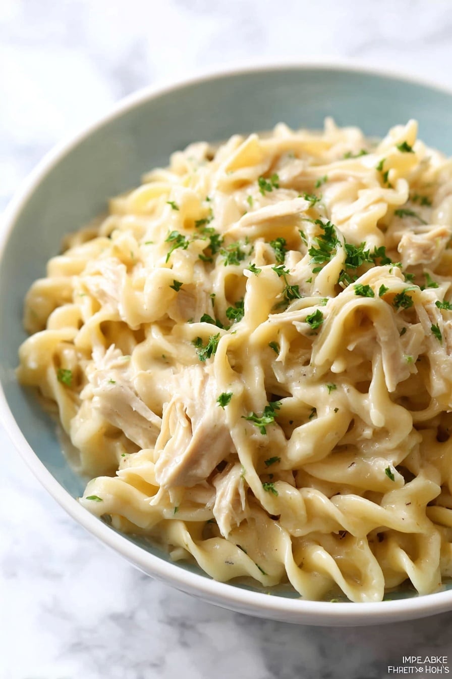 A close-up of a white bowl filled with creamy egg noodles mixed with shredded pieces of light beige chicken. The egg noodles are thick and twisted, coated in a smooth, light cream sauce. Small bits of green herbs are sprinkled on top and some are mixed within the noodles, adding small dots of green color. The bowl is set on a white marbled surface, creating a clean and bright background. Photo taken with an iphone --ar 2:3 --v 7 - Crockpot Chicken and Noodles, easy slow cooker chicken dinner, Creamy chicken noodle casserole, make-ahead comfort food, family-friendly crockpot meal