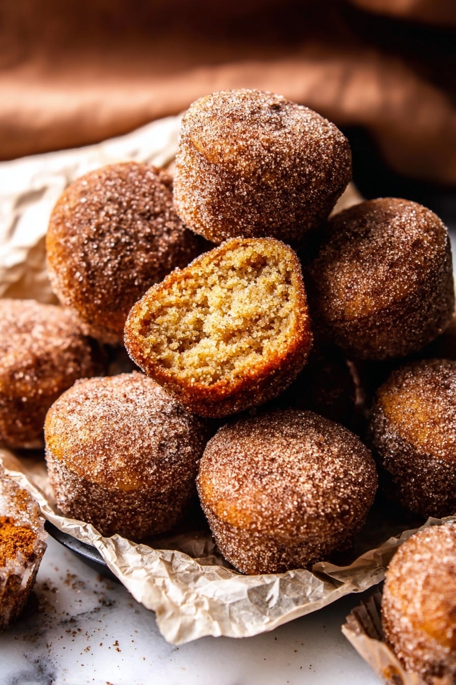 The image shows a pile of small, round doughnut muffins covered in cinnamon sugar on a white marbled surface. There are about ten muffins closely stacked, with one muffin on top showing its inside layer which is soft and light brown with a slightly crumbly texture. The muffins have a rough, grainy outer layer from the sugar coating, and they are resting on crumpled white parchment paper. The scene captures a warm, cozy feeling from the brown and golden hues of the muffins, with some muffins sitting inside a black tray. photo taken with an iphone --ar 2:3 --v 7 - Baked Cinnamon Sugar Apple Cider Doughnut Holes, fall appetizer, cinnamon sugar doughnut holes, baked doughnut holes recipes, cozy fall dessert