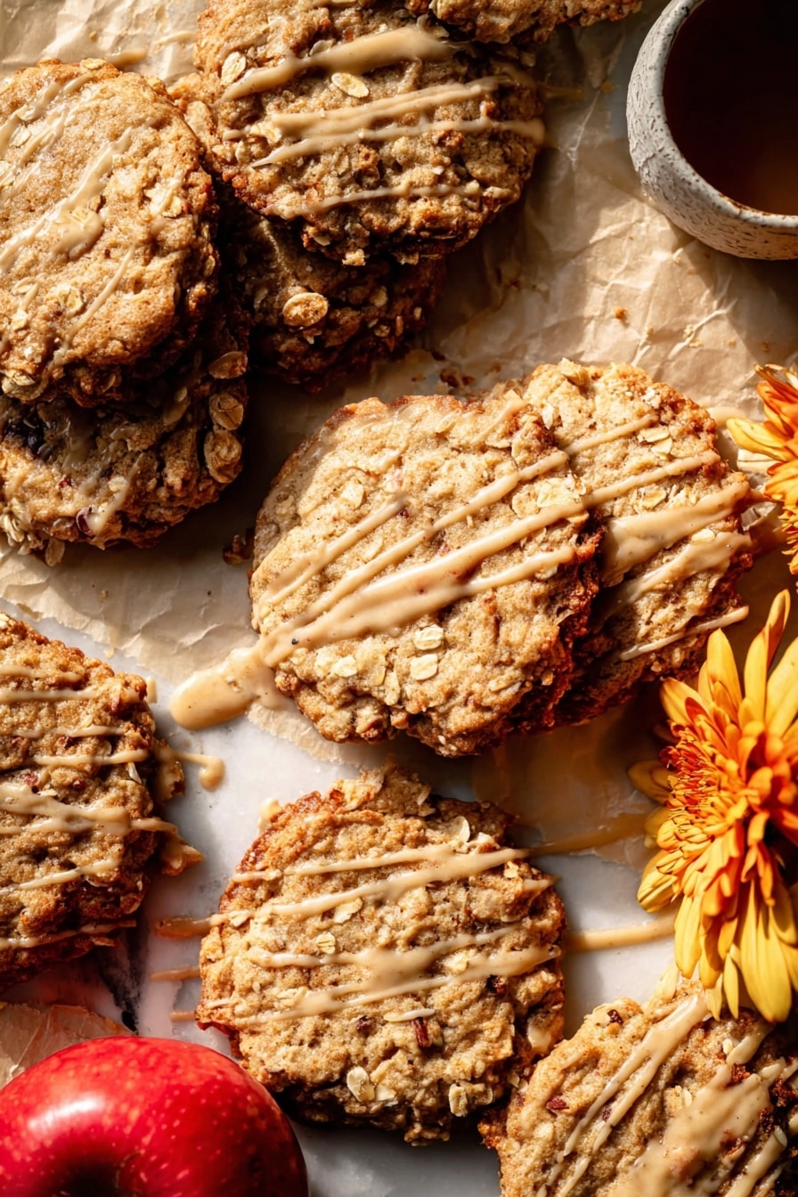 Several oatmeal cookies are spread on light brown parchment paper over a white marbled surface. The cookies are golden brown with a rough texture, showing visible oats and slightly darker edges that look crispy. Each cookie is drizzled with a thin layer of light caramel-colored glaze, creating irregular lines and patches across their tops. In the bottom left corner of the image, two whole red apples with light yellow patches are placed together, adding a fresh contrast to the warm cookie colors. The light source creates soft shadows and highlights, enhancing the cookies’ textures and the glaze's shine. photo taken with an iphone --ar 2:3 --v 7 - Iced Apple Cider Caramel Oatmeal Cookies, apple cider oatmeal cookies, caramel apple cookies, autumn oatmeal cookies, chewy apple cookies