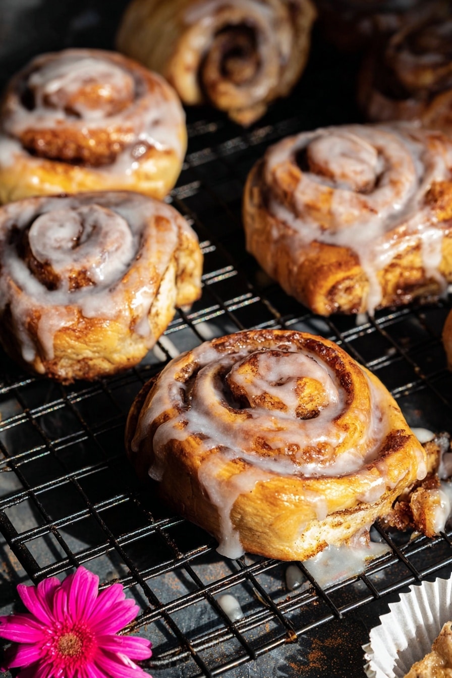 The image shows several cinnamon rolls on a black wire cooling rack with a dark base underneath. Each cinnamon roll has visible spiral layers of golden-brown dough swirled with darker brown cinnamon sugar. The top layers are coated with a shiny white glaze that drips slightly down the sides and onto the rack. The rolls have a soft, fluffy texture with a mix of light yellow and brown tones, and some darker, slightly crisped edges. Near the bottom right, there is a white paper liner with a bright pink flower placed on it. The light creates highlights on the glossy glaze and shadows between the cinnamon rolls, giving the scene depth and warmth. Photo taken with an iphone --ar 2:3 --v 7 - Shortcut Easy Apple Fritters, quick apple fritters, easy apple dessert, fall apple treats, flaky puff pastry apple snacks