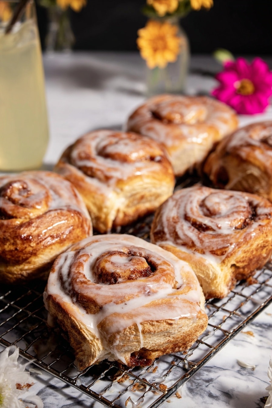 The image shows six cinnamon rolls with a thick white glaze, placed on a wire cooling rack over a white marbled surface. Each cinnamon roll has multiple golden brown layers swirled tightly together with visible cinnamon filling, some layers darker due to caramelized sugar. The glaze drips slightly down the sides, adding a shiny texture on top of the soft, fluffy layers. A white paper liner holds one cinnamon roll in the front, surrounded by a few splashes of glaze on the surface. In the background, a tall glass with a light yellow liquid and a flower in a small white vase add a soft decorative touch, along with a pink flower in a white paper liner near the lower right corner. photo taken with an iphone --ar 2:3 --v 7 - Shortcut Easy Apple Fritters, quick apple fritters, easy apple dessert, fall apple treats, flaky puff pastry apple snacks
