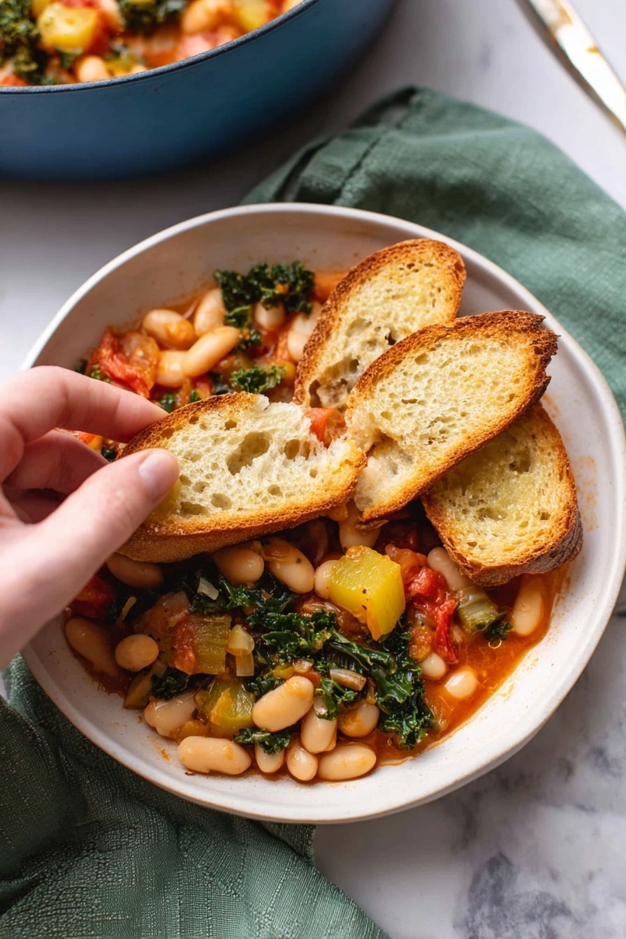 A white bowl filled with a warm stew made of white beans, chunks of yellow squash, wilted green kale, and small red tomato pieces. On top, three slices of toasted baguette bread rest, one slice held by a woman's hand, showing the mix of beans and vegetables piled on it. The stew's sauce is a light red color, and the bread has a golden-brown crust with a porous, soft inside texture. The bowl sits on a green cloth on a white marbled surface, with part of a blue pot filled with more stew blurred in the background. Photo taken with an iphone --ar 2:3 --v 7 - Stewed Great Northern Beans with Kale, Tomatoes, and Herbs, hearty bean and kale stew, healthy vegetarian dinner, wholesome bean recipes, easy bean and greens dish