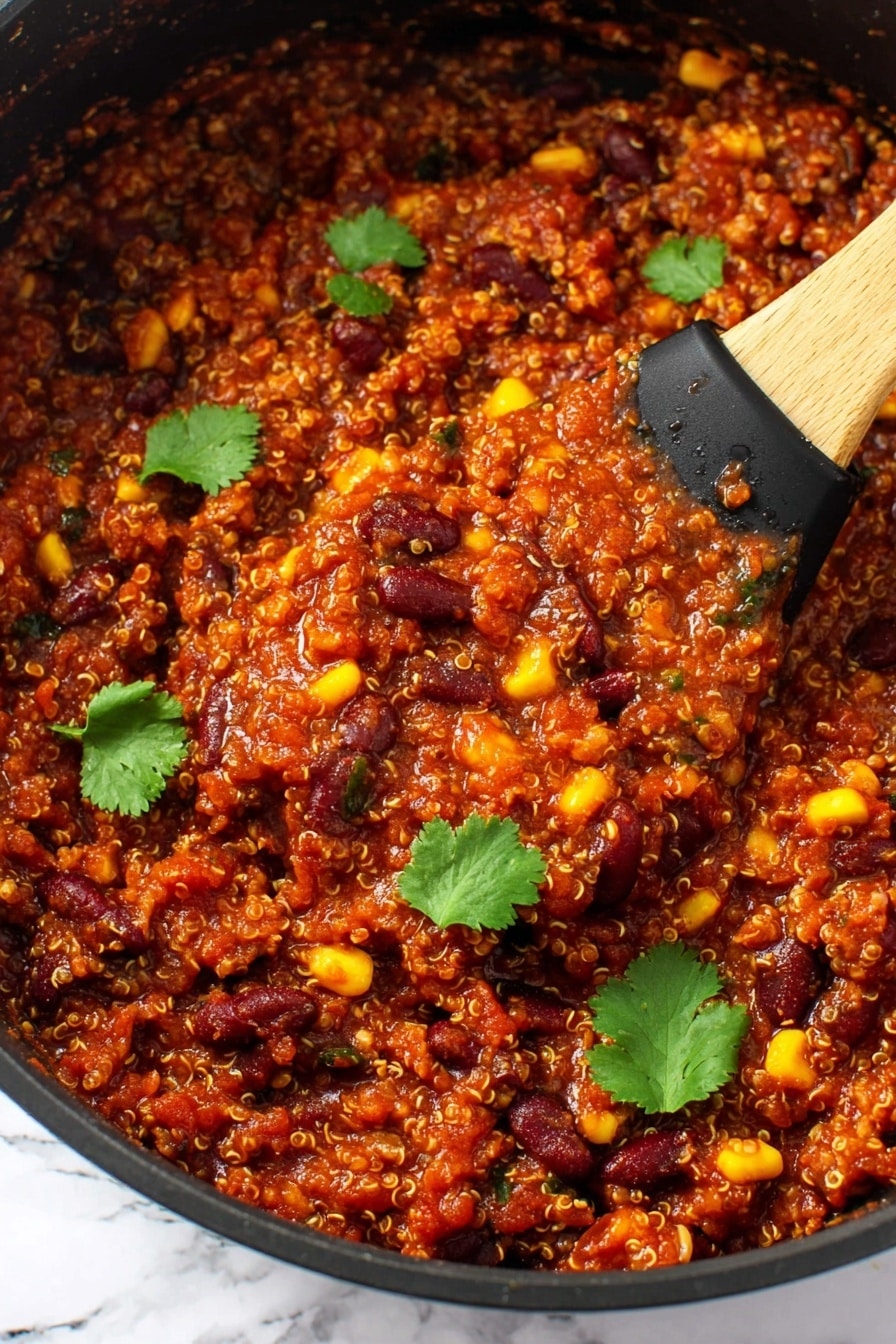 The image shows a thick, rich mixture in a black pan filled with three main layers: grains that look like tiny beige quinoa, dark red kidney beans, and bright yellow corn kernels, all mixed with a deep red tomato-based sauce. The texture is chunky with small bits of vegetables and beans visible throughout. There are several fresh green cilantro leaves scattered on top for contrast. A wooden spoon with a black silicone head is scooping some of the mixture from the pan. The background is a white marbled texture. Photo taken with an iphone --ar 2:3 --v 7 - Slow Cooker Vegan Bean & Quinoa Chili, vegan chili recipe, easy vegan chili, healthy slow cooker dinner, plant-based chili