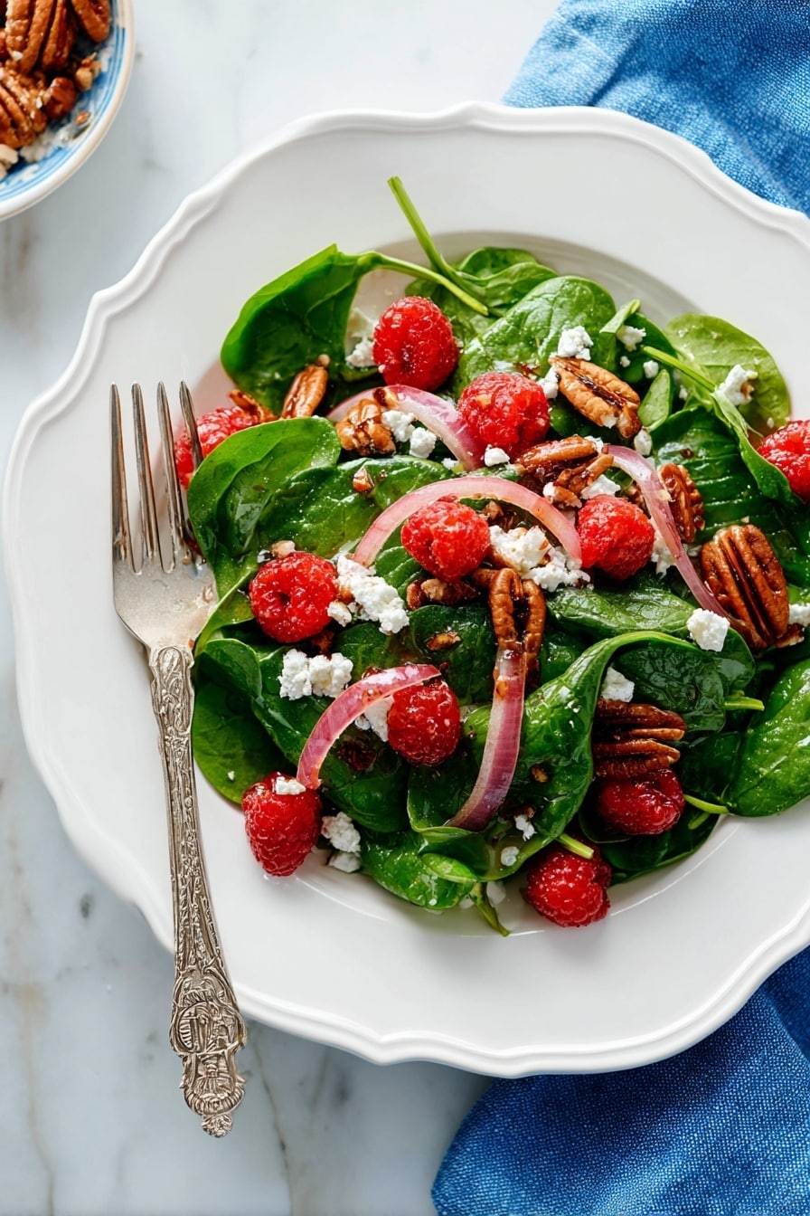 A white scalloped plate holds a fresh salad with three main layers: the base layer has bright green spinach leaves with soft texture, the middle layer has thin slices of pale pink-red onions scattered evenly, and the top layer features bright red raspberries, crumbled white cheese, and brown pecan halves arranged to create color contrast. A silver fork with an ornate handle rests on the left side of the plate, partially on the salad. The plate is placed on a white marbled surface with a blue cloth napkin nearby. Photo taken with an iphone --ar 2:3 --v 7 - Sweet and Tangy Spinach Salad with Goat Cheese, Raspberries, and Toasted Pecans, spinach salad with goat cheese and raspberries, easy fruit and nut salad recipes, healthy berry spinach salad, quick gourmet salad ideas