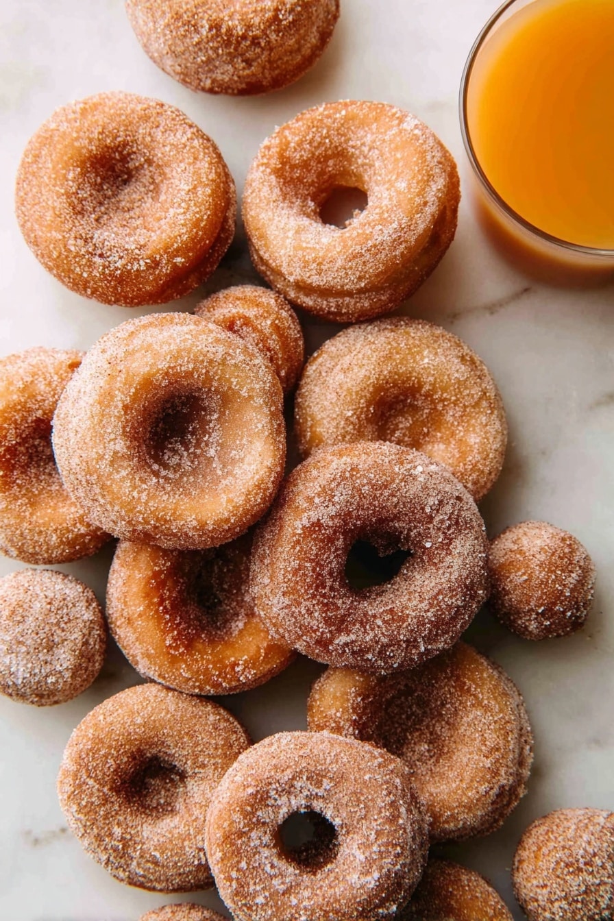 A pile of about a dozen small round donuts with light brown color, coated evenly with granulated sugar and cinnamon, arranged casually on a white marbled surface; two small round donut holes are also visible among the donuts, and at the top right corner, a clear glass of orange-colored juice is partially shown. photo taken with an iphone --ar 2:3 --v 7 - Super Moist Apple Cider Donuts with Warm Spices, apple cider donuts recipe, fall donuts with warm spices, easy apple cider donut recipe, cozy autumn dessert