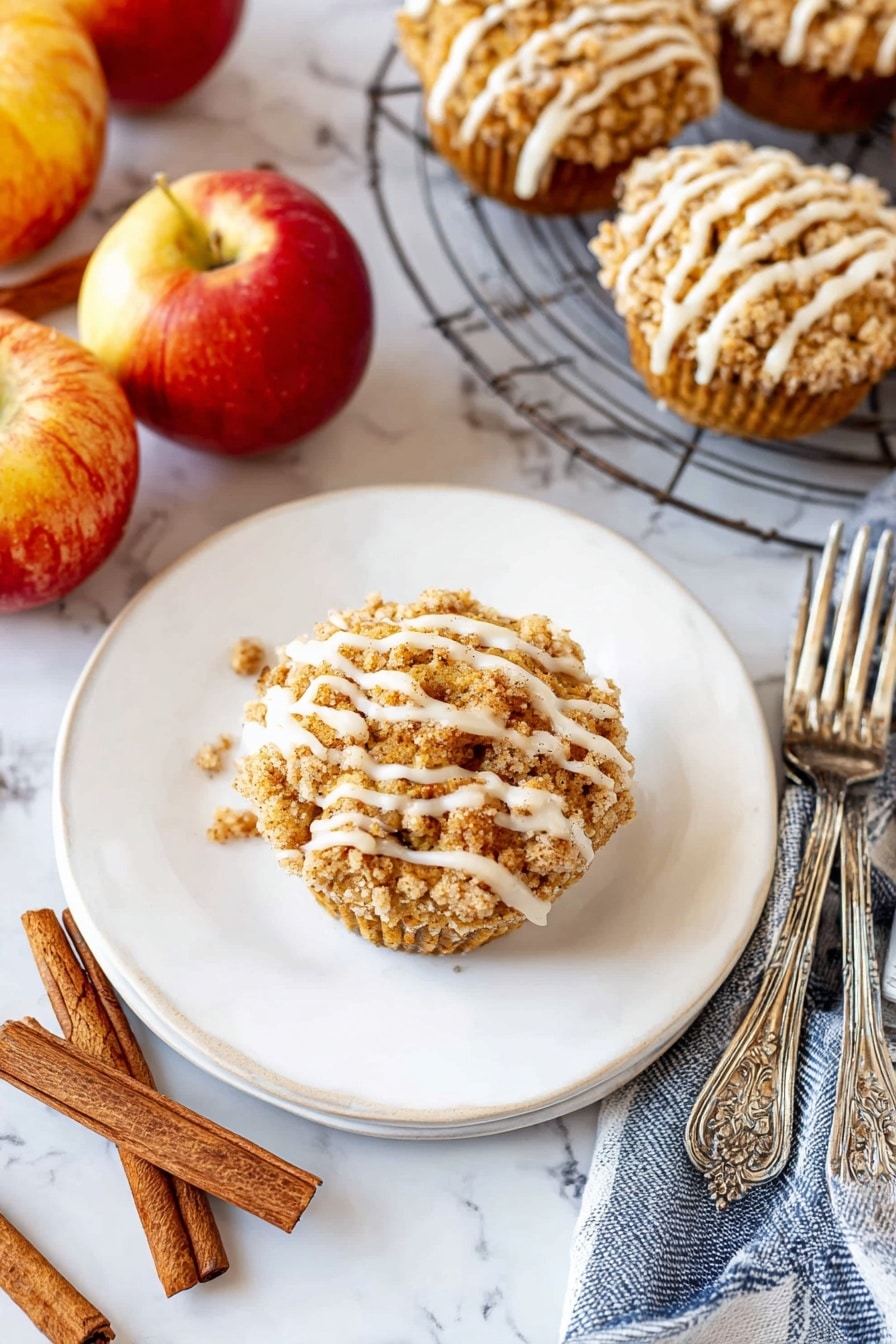 A single round muffin sits in the middle of a white plate, topped with a crumbly golden-brown streusel layer and drizzled with thick white icing in uneven stripes. The plate is on a white marbled surface surrounded by three shiny red and yellow apples, two cinnamon sticks, and a blue and white striped cloth. To the right, a wire cooling rack holds several more muffins with the same crumb topping and white icing drizzle. Two ornate silver forks rest beside the plate. The scene is bright and cozy, with warm autumn colors and textures. photo taken with an iphone --ar 2:3 --v 7 - Cinnamon Apple Muffins with Crumble Topping and Glaze, cinnamon apple muffins, apple spice muffins, homemade apple muffins, easy apple muffin recipes