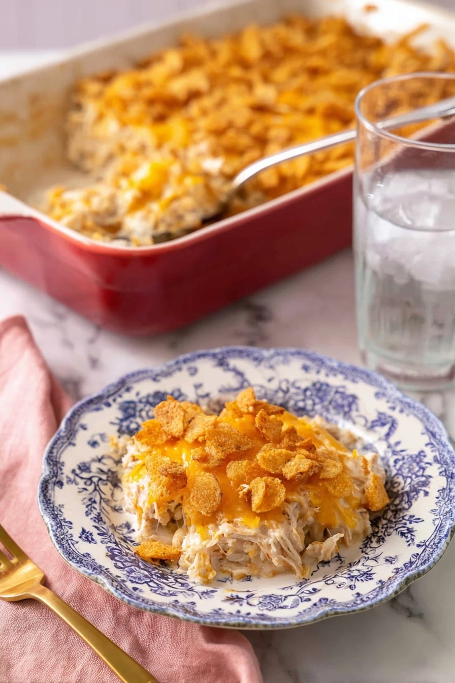 A portion of a creamy casserole is served on a white plate with blue floral patterns, showing three main layers: the bottom layer is shredded white meat mixed with a light creamy sauce, the middle layer has melted orange cheese blended with the meat, and the top layer consists of crushed crispy crackers with more melted cheese giving a golden-brown, crunchy texture. Behind the plate, there is a large red baking dish filled with the same casserole, with a silver serving spoon inside. To the right of the plate is a clear glass of ice water, and to the left is a gold fork resting on a pink cloth napkin, all placed on a white marbled surface. photo taken with an iphone --ar 2:3 --v 7 - Cheesy Chicken Casserole, easy chicken casserole, creamy chicken bake, cheesy chicken dinner, weeknight chicken recipes
