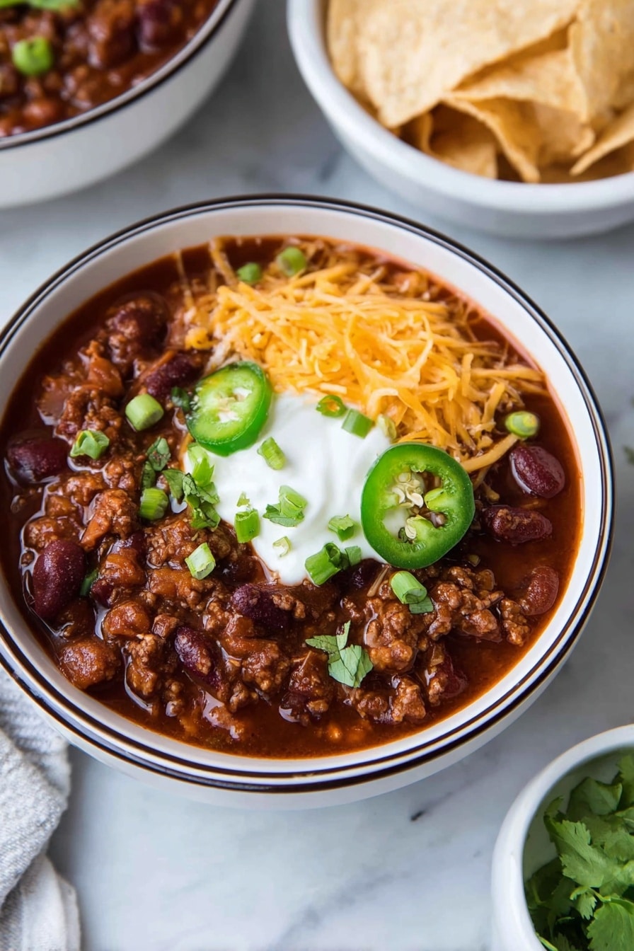 A white bowl with a dark rim holds a thick dark red chili filled with beans and ground meat. On top, there is a layer of shredded orange cheddar cheese on one side, a dollop of white sour cream in the center, sliced green jalapeño rings below, and chopped green onions and cilantro scattered around. In the background, there is a white bowl filled with light-colored tortilla chips on a white marbled surface. Photo taken with an iphone --ar 2:3 --v 7 - Healthy Turkey Chili, healthy chili recipes, turkey chili with beans, low-fat chili dinner, nourishing comfort food