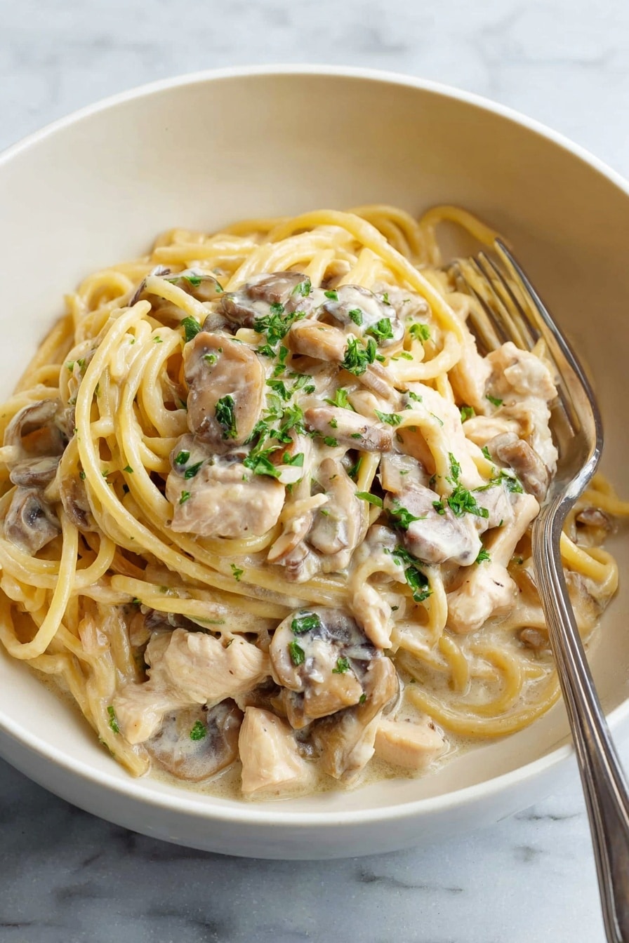 A white bowl holds creamy pasta with three main layers: at the bottom are light brown mushroom pieces coated in sauce, on top are thick, pale yellow spaghetti noodles mixed with tender white chicken pieces, all covered in a smooth, light cream sauce. Small green parsley bits are scattered on top, adding a touch of color. A silver fork rests inside the bowl on the right side. The bowl sits on a white marbled surface. Photo taken with an iphone --ar 2:3 --v 7 - Creamy Chicken Tetrazzini with Mushrooms, Chicken Mushroom Pasta Bake, Easy Creamy Chicken Casserole, Comforting Chicken and Mushroom Dinner, Baked Chicken Pasta with Mushrooms