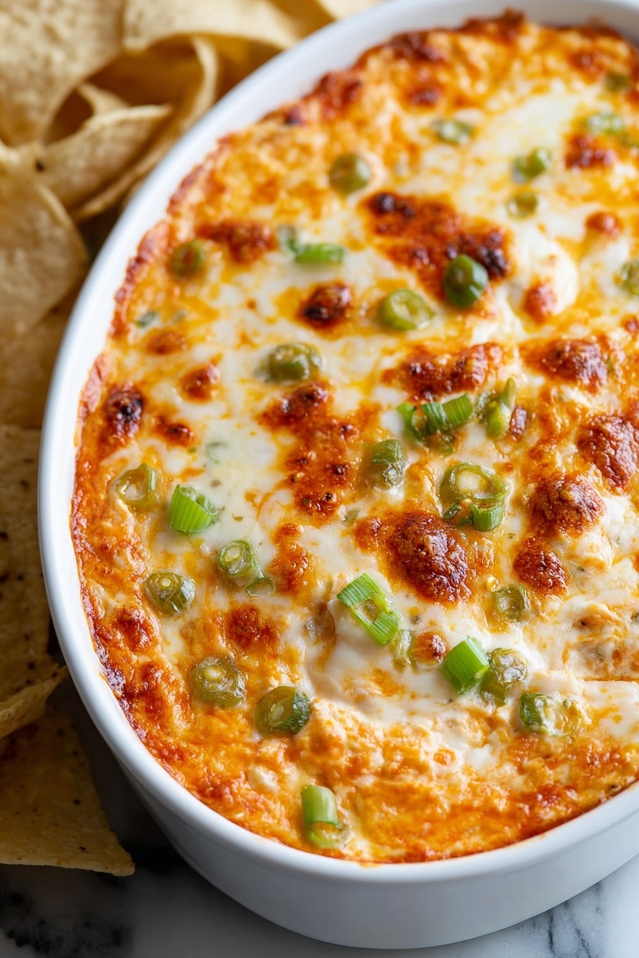 The image shows a close-up of a white oval dish filled with a baked cheesy dip. The top layer is golden brown melted cheese with small browned spots and melted orange cheese mixed in. Scattered on the surface are pieces of green onion adding a fresh green color contrast. The dish is placed on a white marbled surface, and in the background, there are some light and dark tortilla chips slightly out of focus. The melted cheese has a bubbly, slightly crispy texture on top photo taken with an iphone --ar 2:3 --v 7 - Buffalo Chicken Dip, spicy chicken dip, cheesy chicken appetizer, game day dip, crowd-pleasing appetizer