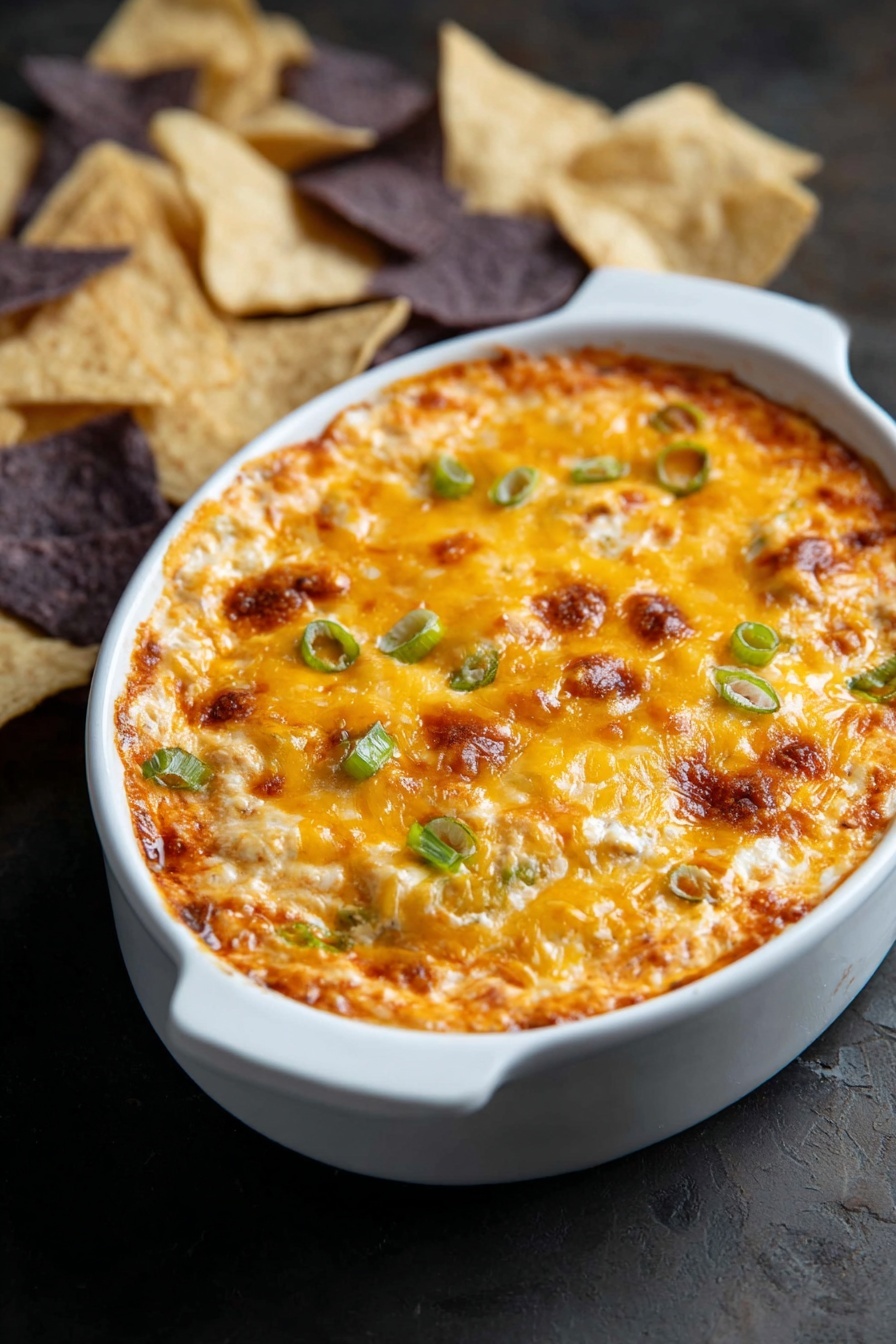 The image shows a white oval dish filled with a baked dip that has a golden-brown melted cheese layer on top with some browned spots. The cheese surface is dotted with small green onion pieces, giving a pop of green color. The dip looks creamy with slightly uneven texture, suggesting layers of ingredients underneath the cheese. The dish sits on a dark surface, and there are light and dark tortilla chips placed around the dish in the background. photo taken with an iphone --ar 2:3 --v 7 - Buffalo Chicken Dip, spicy chicken dip, cheesy chicken appetizer, game day dip, crowd-pleasing appetizer