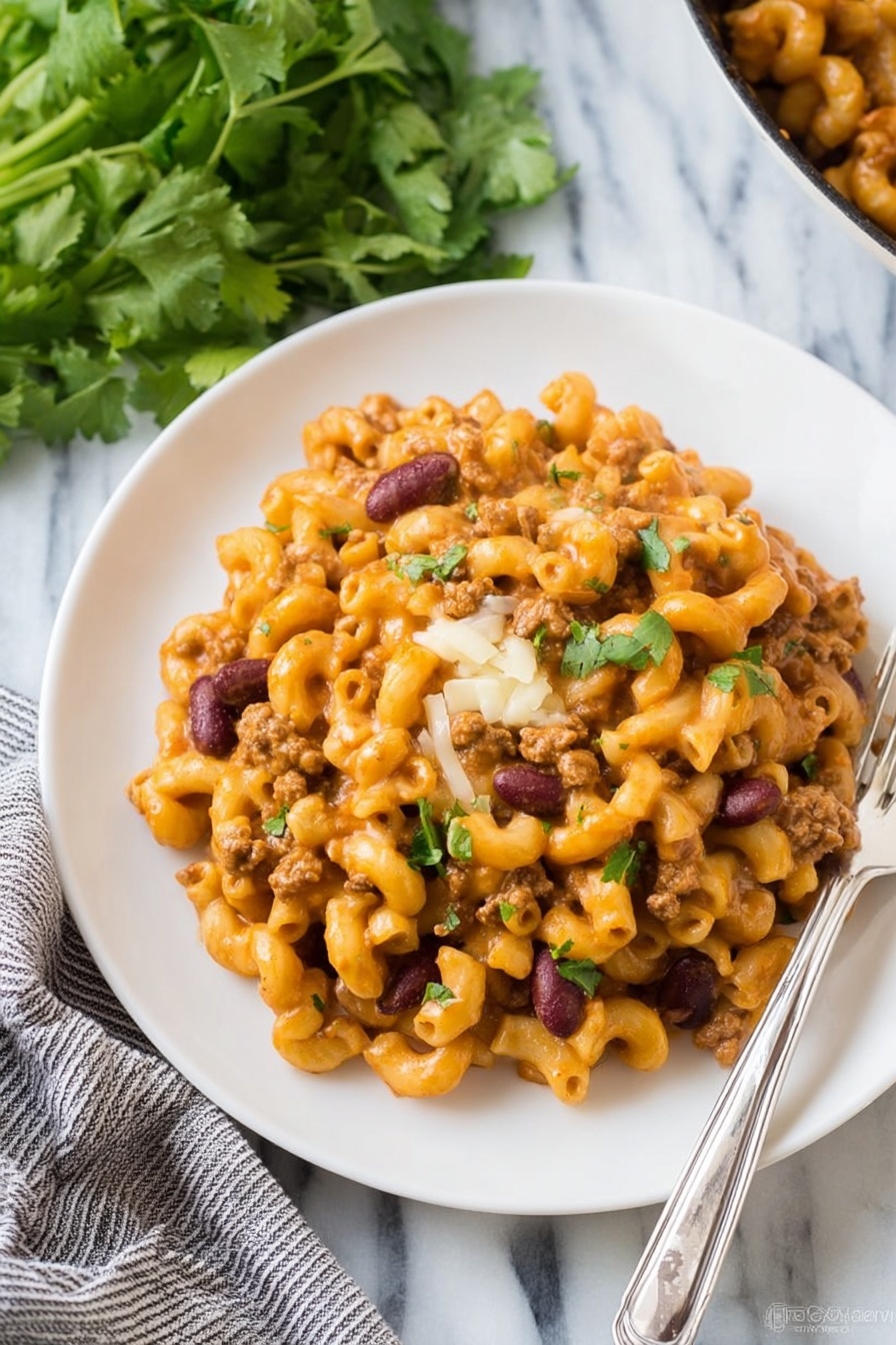 The dish shows a single layer of cooked elbow macaroni mixed with a creamy orange sauce, ground meat, and dark red kidney beans, all placed on a round white plate. The macaroni and sauce mix looks thick and slightly glossy, with small bits of green herbs sprinkled on top, adding a touch of color. A small amount of melted white cheese sits near the center, blending with the sauce. The plate rests on a white marbled surface with a silver fork beside it on the right, and in the background, there is a bunch of fresh green leafy herbs and a gray and white striped cloth. Photo taken with an iphone --ar 2:3 --v 7 - Turkey Chili Mac and Cheese, easy chili mac recipe, hearty dinner ideas, quick weeknight meals, protein-packed comfort food