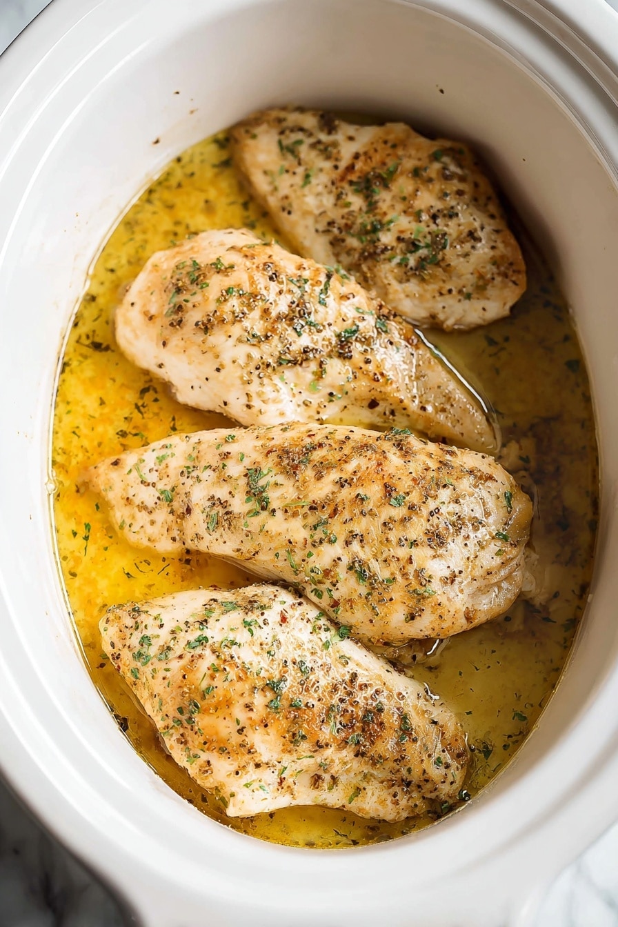 The image shows four pieces of cooked chicken placed side by side inside a white slow cooker. Each piece is light golden brown with some darker brown spots from cooking and sprinkled with green herbs and black pepper. The chicken sits in a shallow puddle of yellowish oil or broth that gives a slight shine on the surface. The inside of the slow cooker is white and smooth, contrasting with the warm tones of the chicken and liquid. The setting features a white marbled background. photo taken with an iphone --ar 2:3 --v 7 - Slow Cooker Juicy Seasoned Chicken Breast, easy slow cooker chicken recipes, tender chicken breast in slow cooker, flavorful chicken breast dinner, healthy slow cooker chicken