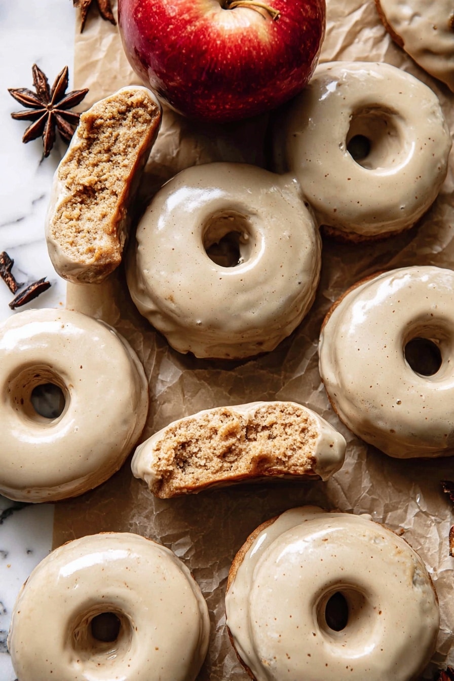 The image shows a group of doughnuts with a smooth, light beige glaze covering their top surfaces. Each doughnut has one round hole in the center and a slightly rough, crumbly texture inside where some are broken or bitten. The doughnuts are laid on a brown paper surface with pieces of white parchment paper scattered around. There is a shiny red apple near the center top of the image and a dark brown star anise near the upper left side. The background is a white marbled texture. photo taken with an iphone --ar 2:3 --v 7 - Baked Apple Cider Doughnuts with Cinnamon Maple Glaze, baked apple cider doughnuts, fall cinnamon glazed doughnuts, homemade apple cider donuts, easy baked doughnut recipe