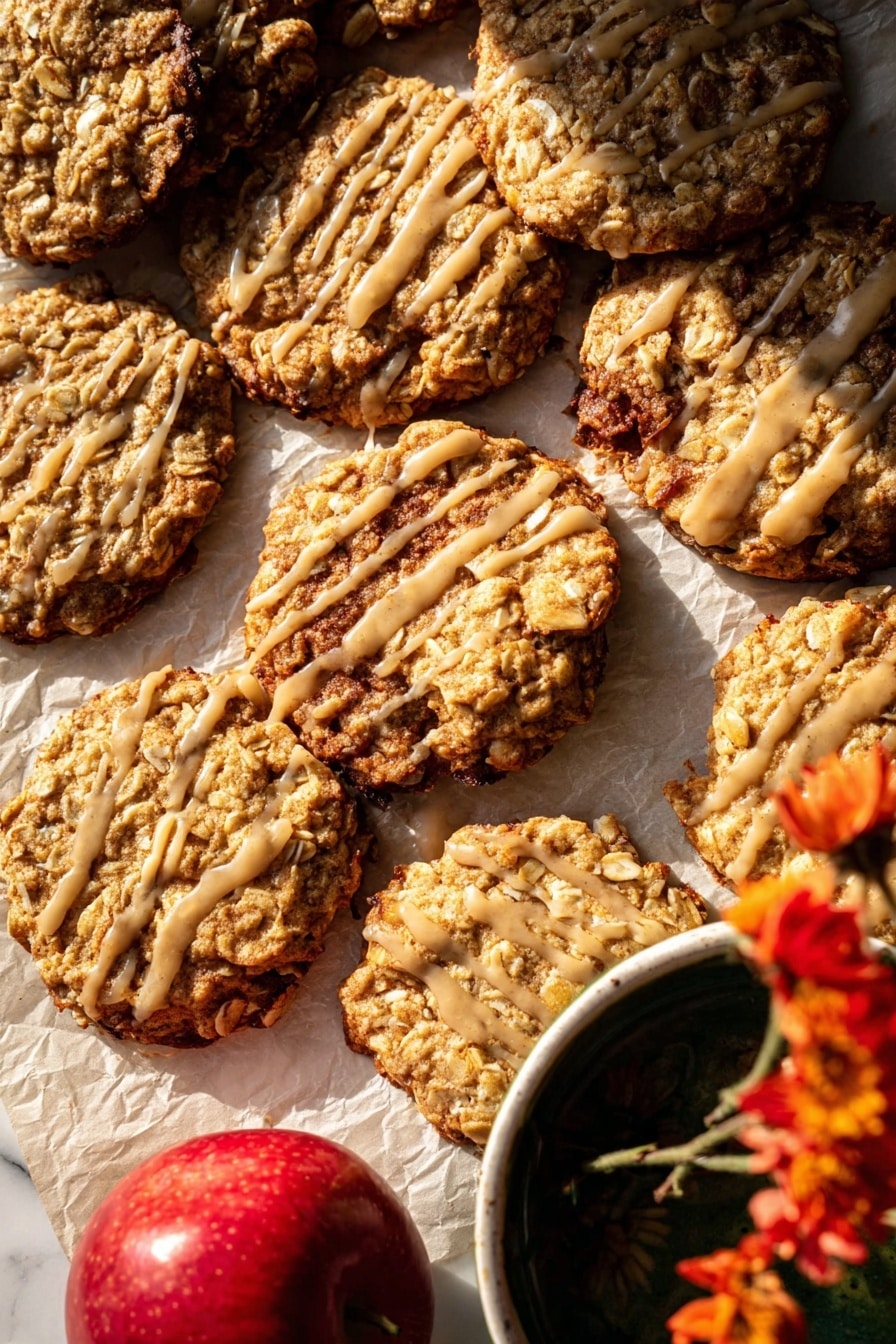 The image shows a close-up of several oatmeal cookies with a slightly rough texture from oats and nuts, arranged unevenly on parchment paper over a white marbled surface. Some cookies are stacked, while others lay flat, revealing their golden-brown baked edges with a few darker spots. A few cookies are drizzled with a light beige glaze in thin, irregular lines, adding shine and contrast to the surface. A bright red apple is partially visible in the bottom left, and the corner of a white bowl with a dark liquid and a yellow flower is seen in the top right, adding color variety. The warm lighting highlights the cookies' texture and the glaze’s glossiness. photo taken with an iphone --ar 2:3 --v 7 - Iced Apple Cider Caramel Oatmeal Cookies, apple cider oatmeal cookies, caramel apple cookies, autumn oatmeal cookies, chewy apple cookies