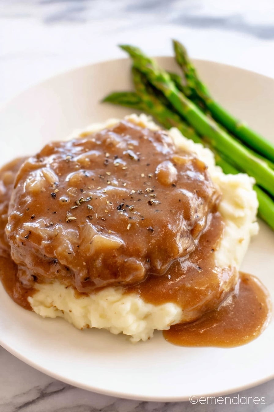 The image shows a white plate with three layers of food. The bottom layer is creamy mashed potatoes, which look soft and smooth in a pale cream color. On top of the mashed potatoes is a thick piece of cooked meat covered generously with a brown gravy sauce that has small onion bits and black pepper sprinkled on top. To the side of the meat, there is a small bunch of cooked green asparagus spears that add a fresh green color. The white plate sits on a white marbled surface. Photo taken with an iphone --ar 2:3 --v 7 - Crock Pot Cube Steak with Creamy Mushroom Gravy, slow cooker cube steak, easy beef dinner, tender beef recipes, mushroom gravy dishes