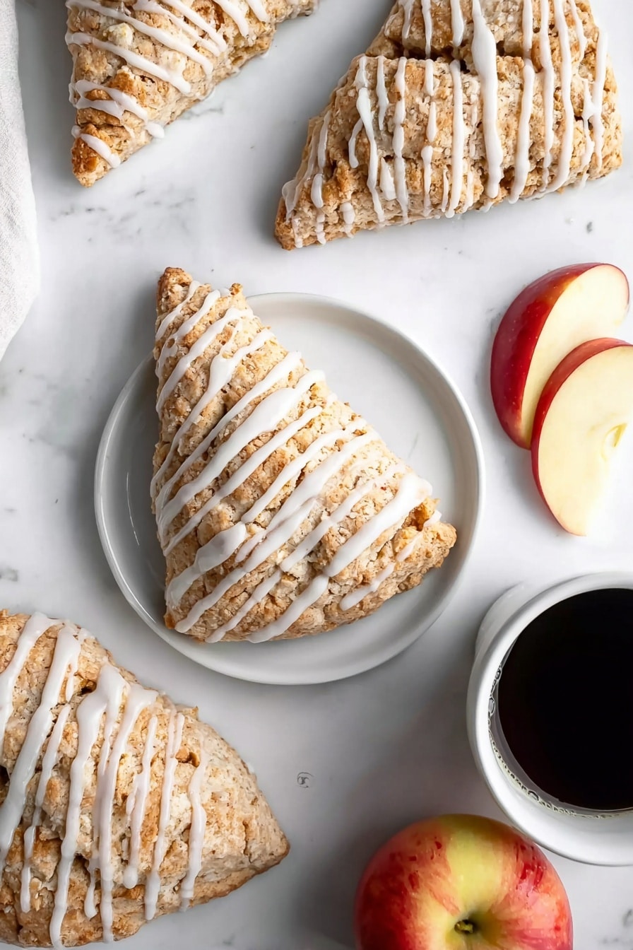 Several triangular scones are arranged on a white marbled surface, each covered in a light brown, crumbly texture with white icing drizzled in even stripes across the top. One scone sits on a simple white plate centered in the image, accompanied by two slices of red and yellow apple on the right side of the plate. Nearby, a whole apple with red and yellow skin is partly visible at the bottom right. In the upper right corner, a white cup filled with black coffee adds contrast. The scene is bright and clean, highlighting the soft texture of the scones and fresh fruit. photo taken with an iphone --ar 2:3 --v 7 - Easy Apple Scones with Cinnamon Glaze, apple scones recipe, fall scones with cinnamon glaze, homemade apple scones, cozy breakfast scones