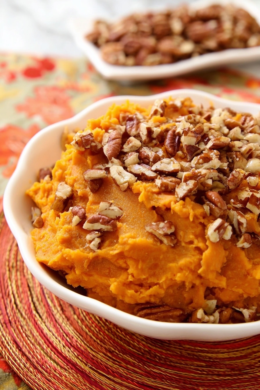 A white scalloped bowl holds a single layer of smooth, bright orange mashed sweet potatoes topped generously with a layer of chopped pecans, which add a textured, brown, and crunchy contrast. In the background, a white dish contains more pecan pieces, slightly out of focus. The bowl and pecans rest on a woven, multi-colored orange and red placemat, with a shiny silver spoon placed nearby. The scene is set on a white marbled surface. Photo taken with an iphone --ar 2:3 --v 7 - Slow Cooker Sweet Potato Casserole with Maple and Toasted Pecans, sweet potato casserole, slow cooker sweet potato dish, holiday sweet potato recipe, easy Thanksgiving side