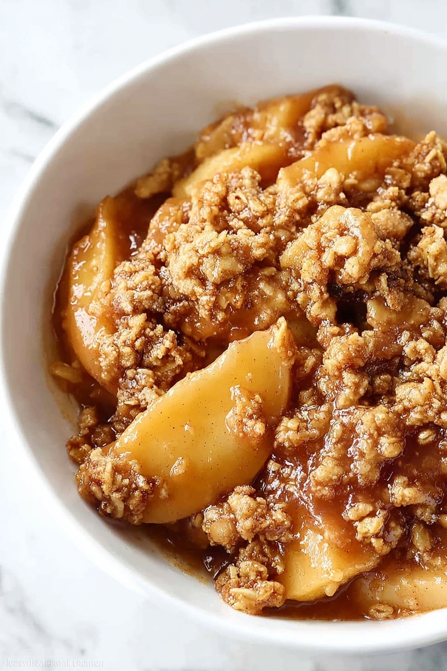 A white bowl holds a warm apple dessert with three distinct layers: the bottom layer has soft, golden-brown apple slices coated in syrupy juices; above this is a crumbly, textured topping made of oats and brown sugar that looks crunchy and slightly toasted; the top layer is a smooth, creamy white scoop of melted vanilla ice cream that softly spreads over the crumble. A silver spoon rests in the bowl, partially visible at the back, all placed on a white marbled surface. Photo taken with an iphone --ar 2:3 --v 7 - Healthy Apple Crisp, healthy apple crisp, healthy apple crisp dessert, wholesome apple crisp recipe, nutritious apple crisp