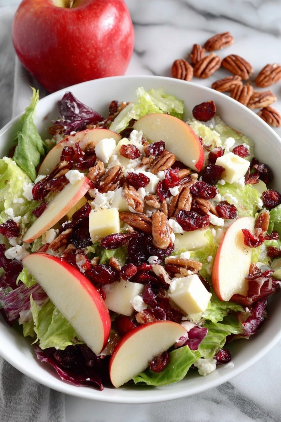 A white bowl filled with a fresh salad sits on a white marbled surface next to a red and yellow apple and a white towel with red stripes. The salad has a base layer of green leafy lettuce, topped with chunks of red and yellow apple pieces. Scattered over the apples are dried red cranberries and brown pecan nuts. The top layer is sprinkled with white crumbled cheese, adding a soft texture contrast to the crunchy ingredients. The photo taken with an iphone --ar 2:3 --v 7 - Honeycrisp Harvest Salad, fall salad with apples, apple and bacon salad, Autumn fruit salad, easy apple salad recipe