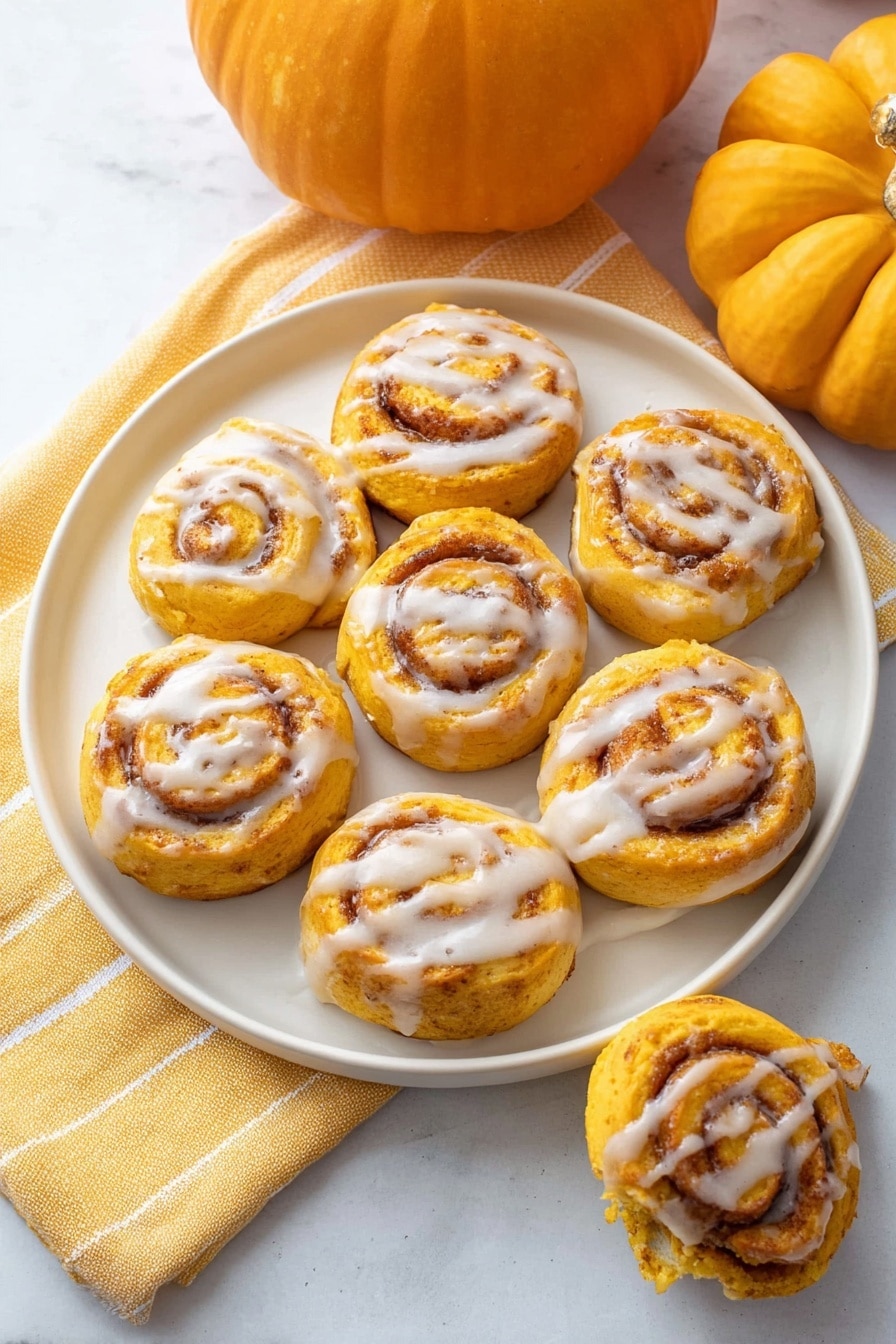 A round white plate holds nine small pumpkin-colored cinnamon rolls arranged in a loose circle. Each roll has light orange and brown swirls visible from the cinnamon filling and is topped with a thin drizzle of white icing, creating a shiny contrast. The plate sits on a soft yellow towel with a white line pattern, placed on a white marbled surface. Two whole pumpkins, one larger and one smaller, sit in the background, adding an autumn feel to the scene. One additional cinnamon roll is partially seen on the bottom right, held by a woman's hand, showing the flaky texture inside. Photo taken with an iphone --ar 2:3 --v 7 - Quick Pumpkin Cinnamon Rolls, pumpkin cinnamon rolls, easy fall breakfast, no yeast cinnamon rolls, quick pumpkin dessert