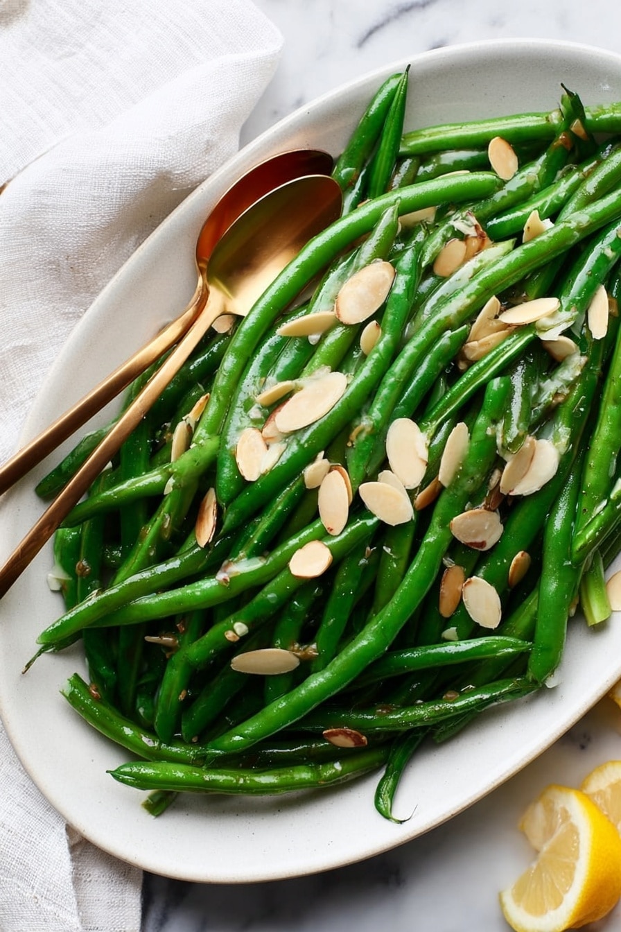 A white oval plate filled with bright green, shiny cooked green beans, arranged in a loose pile covering the plate. On top of the beans are thin, light tan almond slices scattered evenly, adding texture and contrast. Two small gold spoons rest on the beans, slightly overlapping, showing off the glossy surface of the veggies. The plate is placed on a white marbled surface, with a white cloth partially visible on the left side and a small piece of lemon on the right edge. The scene is well-lit, emphasizing the freshness and color of the dish. photo taken with an iphone --ar 2:3 --v 7 - Green Beans Almondine with Toasted Almonds, green beans almondine, lemon green bean side dish, almond-sautéed green beans, elegant vegetable side dish