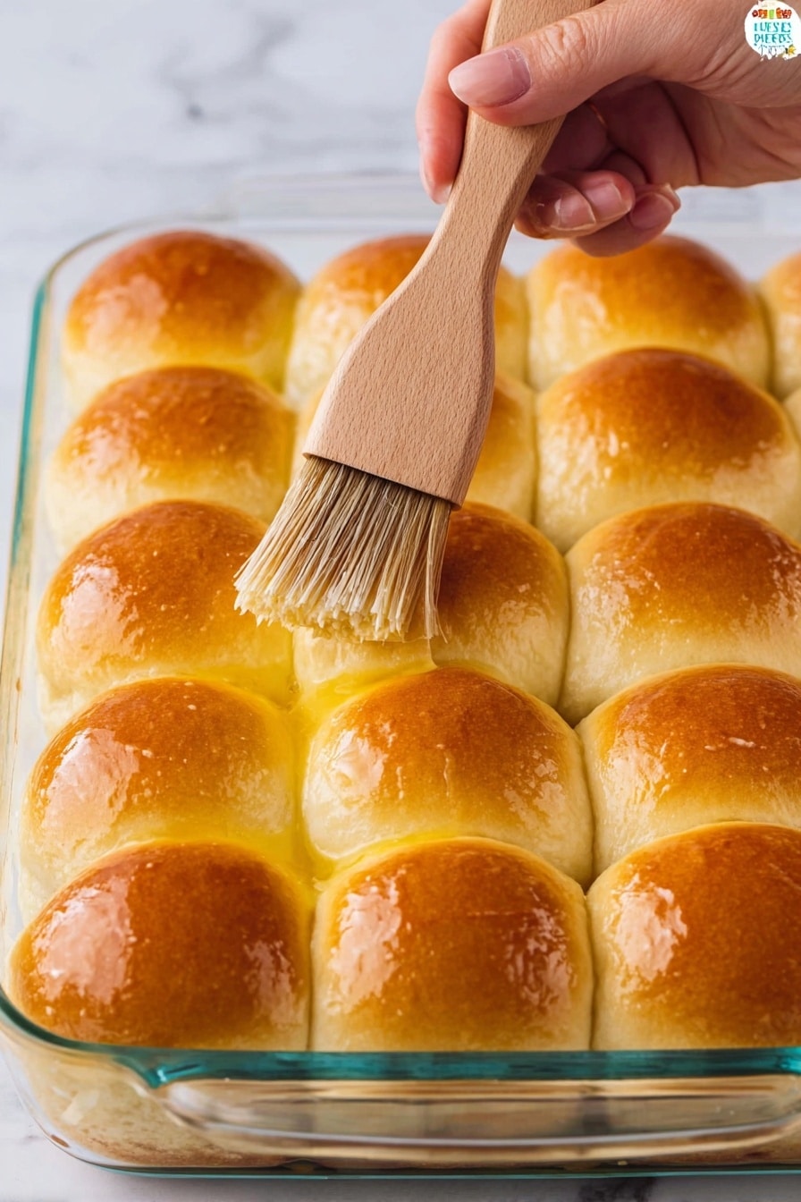 A glass baking dish holds a layer of 12 evenly arranged soft dinner rolls, each roll golden brown on top and shiny from being brushed with melted butter. The rolls sit closely together in a grid pattern with a soft, slightly fluffy texture. A woman's hand holds a light wooden brush with brown bristles, applying butter to the top of one roll. The background is a white marbled surface. photo taken with an iphone --ar 2:3 --v 7 - Homemade Fluffy Dinner Rolls, fluffy dinner rolls recipe, soft buttery dinner rolls, easy homemade dinner rolls, bakery-style dinner rolls