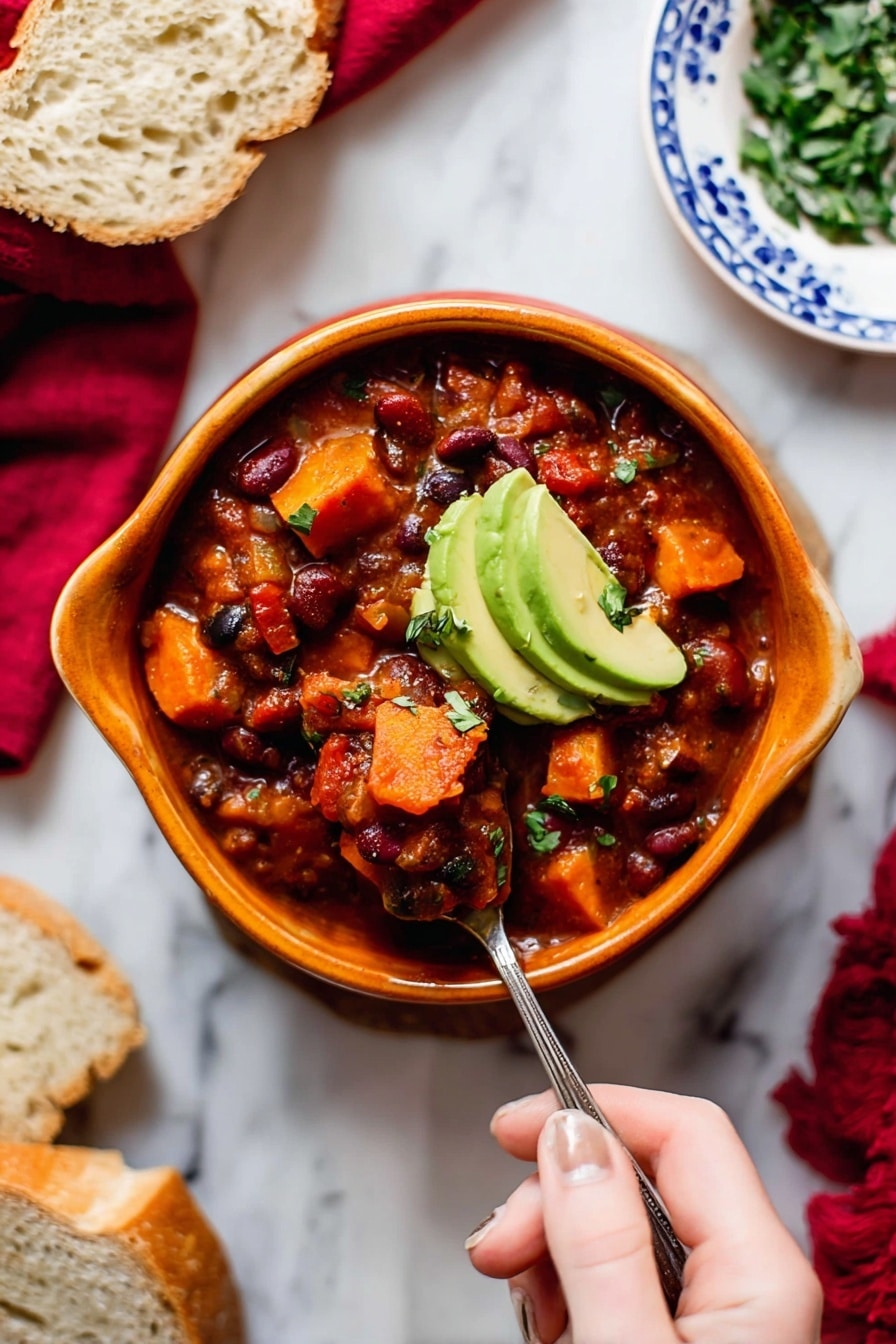 A bowl of thick chili with visible layers of chunky tomato sauce, red and black beans, and soft, cubed orange sweet potatoes, topped with two slices of green avocado. The chili is served in a curved, orange bowl on a white marbled surface, with a woman's hand holding a spoon scooping into the chili. Surrounding the bowl are slices of bread on a white plate with blue patterns, a small bowl of chopped green herbs, and a red cloth in the background. Photo taken with an iphone --ar 2:3 --v 7 - Vegetarian Pumpkin Chili, Healthy Pumpkin Chili, Cozy Vegetarian Dinner, Fall Vegetarian Recipes, Hearty Pumpkin Chili