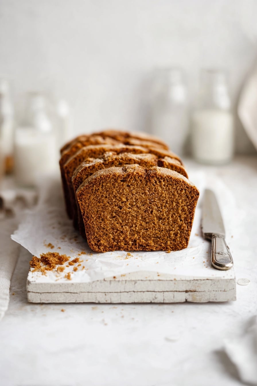 The image shows a loaf of brown bread sliced into six pieces, placed on white parchment paper on a rustic white wooden board. The bread has a golden-brown crust with a slightly rough texture and a dense, soft interior with small air holes. There are some crumbs scattered around the bread on the white marbled surface. To the upper right of the bread, there is a silver knife resting on top of a folded piece of white paper with text, and in the upper left, a clear empty glass and a white container are partially visible. Photo taken with an iphone --ar 2:3 --v 7 - Pumpkin Bread, Pumpkin Bread for Fall, Easy Pumpkin Bread, Moist Pumpkin Bread, Fall Pumpkin Baking