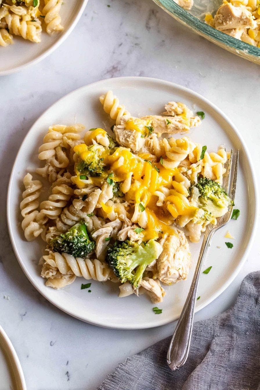 A white plate on a white marbled surface holds a serving of rotini pasta mixed with chunks of cooked chicken, bright green broccoli florets, and melted yellow cheese drizzled unevenly over the top. The pasta spirals are pale cream and slightly glossy, nestled closely with tender pieces of chicken and small green broccoli heads, all lightly sprinkled with small bits of fresh green herbs. A silver fork rests partially on the right edge of the plate, with its tines touching some pasta. There is a gray cloth napkin in the lower right corner and a partial view of another white plate and a glass baking dish in the background. Photo taken with an iphone --ar 2:3 --v 7 - Chicken Pasta Casserole with Broccoli, easy chicken pasta bake, cheesy chicken casserole, healthy broccoli pasta dish, family-friendly comfort food