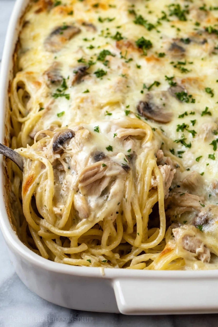 This dish shows a close-up view of a baked pasta casserole in a white ceramic dish. The casserole consists of creamy spaghetti mixed with pieces of light brown cooked meat and sliced mushrooms spread throughout. The top layer is covered with a smooth, melted white cheese sauce sprinkled with small bits of green herbs. A spoon is scooping some of the creamy pasta from the dish, showing the mixed layers of noodles, meat, and mushrooms inside. The dish is placed on a white marbled surface. photo taken with an iphone --ar 2:3 --v 7 - Creamy Chicken Tetrazzini with Mushrooms, Chicken Mushroom Pasta Bake, Easy Creamy Chicken Casserole, Comforting Chicken and Mushroom Dinner, Baked Chicken Pasta with Mushrooms