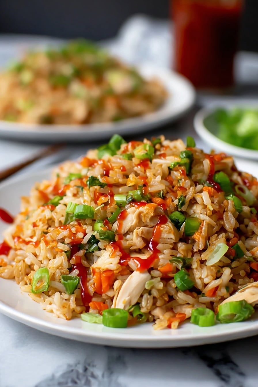 A close-up view of a white plate filled with fried rice, showing layers of light brown rice mixed with small pieces of white chicken, bright green chopped spring onions, and small orange-red peppers scattered throughout. The rice is lightly coated with red sauce drizzled on top, adding a glossy texture. In the background, there is a blurred white plate with more fried rice, placed on a white marbled surface. The overall colors are warm with green, red, and white contrasts. Photo taken with an iphone --ar 2:3 --v 7 - Quick Chicken Fried Rice, easy chicken fried rice, fast fried rice recipe, beginner-friendly fried rice, leftover chicken dinner