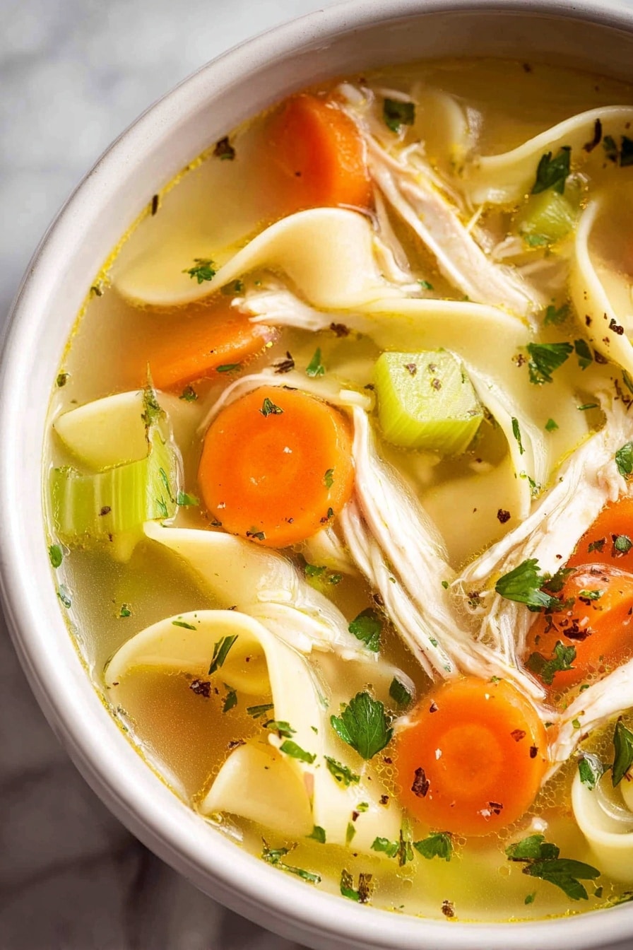 A white bowl filled with clear golden broth containing long, flat yellow noodles, small orange carrot cubes, and green celery pieces. Shredded light beige chicken is spread evenly throughout, with flecks of green herbs and black pepper for extra color. A single dark brown bay leaf is visible near the bottom, and a silver spoon is partially submerged in the soup. The bowl rests on a white marbled surface with a small white cup of chopped green herbs and another white cup with a white creamy substance in the background, photo taken with an iphone --ar 2:3 --v 7 - Homemade Chicken Noodle Soup from Scratch, chicken noodle soup from scratch, homemade chicken soup, hearty chicken noodle recipe, easy homemade chicken soup