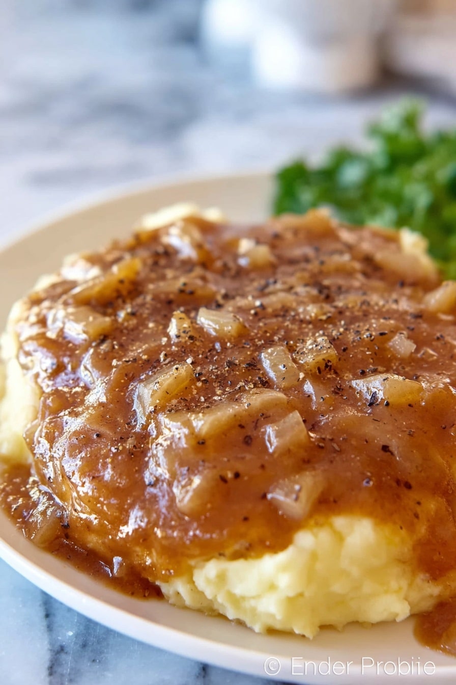 A white plate with mashed potatoes as the base layer, creamy and light yellow in color with a smooth texture, topped with a thick brown gravy that has small diced onion pieces spread evenly across the surface. The gravy covers a large piece of meat underneath, sprinkled with black pepper flakes. The background shows an out-of-focus white marbled surface and some green garnish in the back. Photo taken with an iphone --ar 2:3 --v 7 - Crock Pot Cube Steak with Creamy Mushroom Gravy, slow cooker cube steak, easy beef dinner, tender beef recipes, mushroom gravy dishes