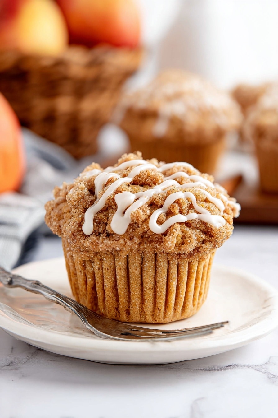 A single muffin sits centered on a white plate with a silver fork resting next to it. The muffin has a light brown, soft textured base with a crumbly streusel topping in a darker brown shade. White icing drizzle runs in thin lines over the streusel, adding contrast. The plate and muffin are placed on a white marbled surface. In the background, blurred muffins and a basket of apples add warm tones but stay out of focus. Photo taken with an iphone --ar 2:3 --v 7 - Cinnamon Apple Muffins with Crumble Topping and Glaze, cinnamon apple muffins, apple spice muffins, homemade apple muffins, easy apple muffin recipes