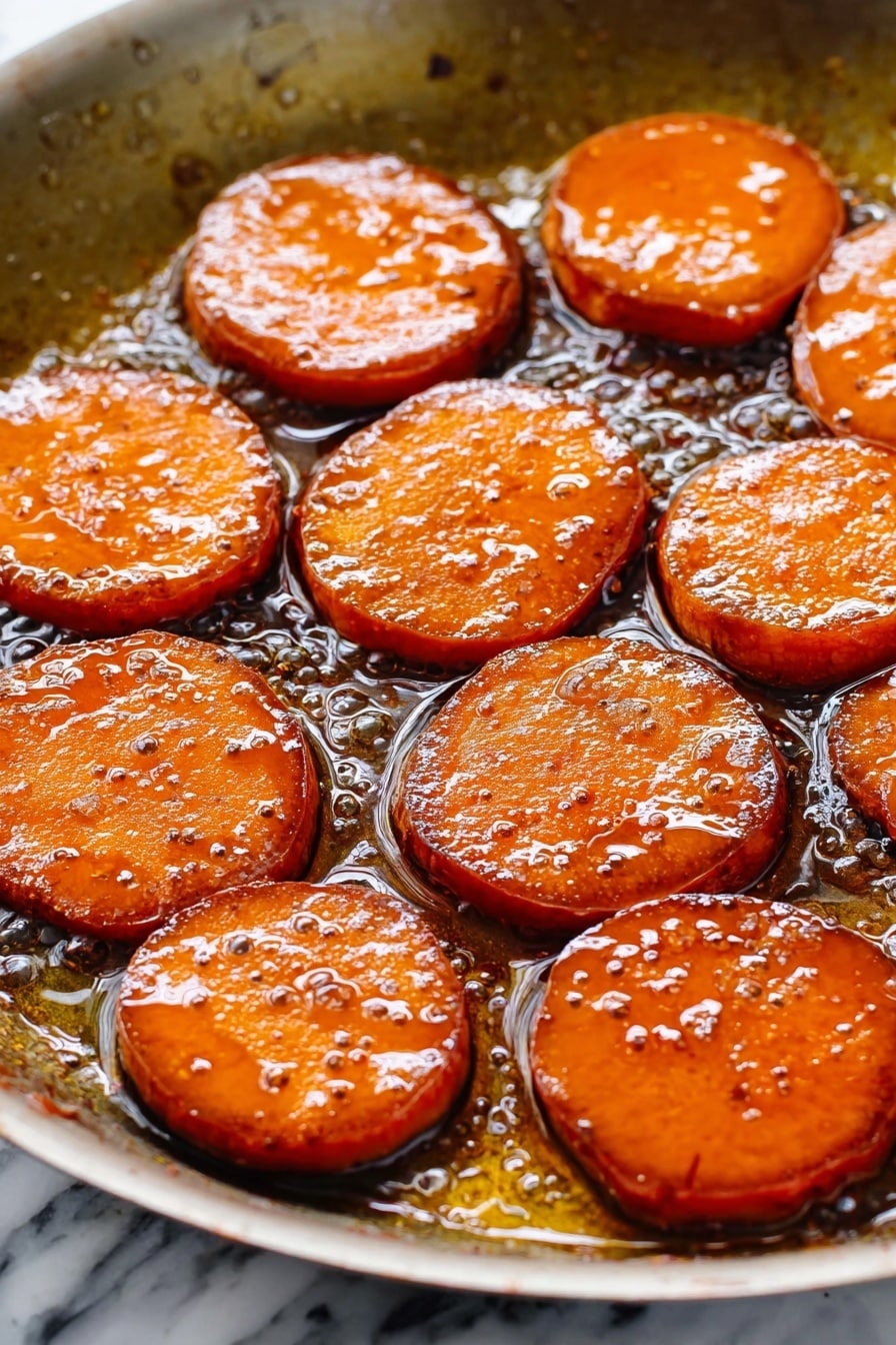 The image shows a white oval dish filled with thick, round slices of cooked sweet potatoes. The sweet potato slices are covered in a shiny, dark caramel-like sauce that makes them look glossy and soft. The slices are arranged closely together, slightly overlapping, and their orange color is rich and warm, enhanced by the dark syrup. The sauce pools around the edges of the slices, filling the bottom of the dish, and the texture looks smooth and sticky. The setting is on a white marbled surface. photo taken with an iphone --ar 2:3 --v 7 - Candied Sweet Potatoes with Bourbon Glaze, bourbon sweet potato side dish, holiday sweet potato recipes, caramelized sweet potatoes with bourbon, easy bourbon glazed sweet potatoes