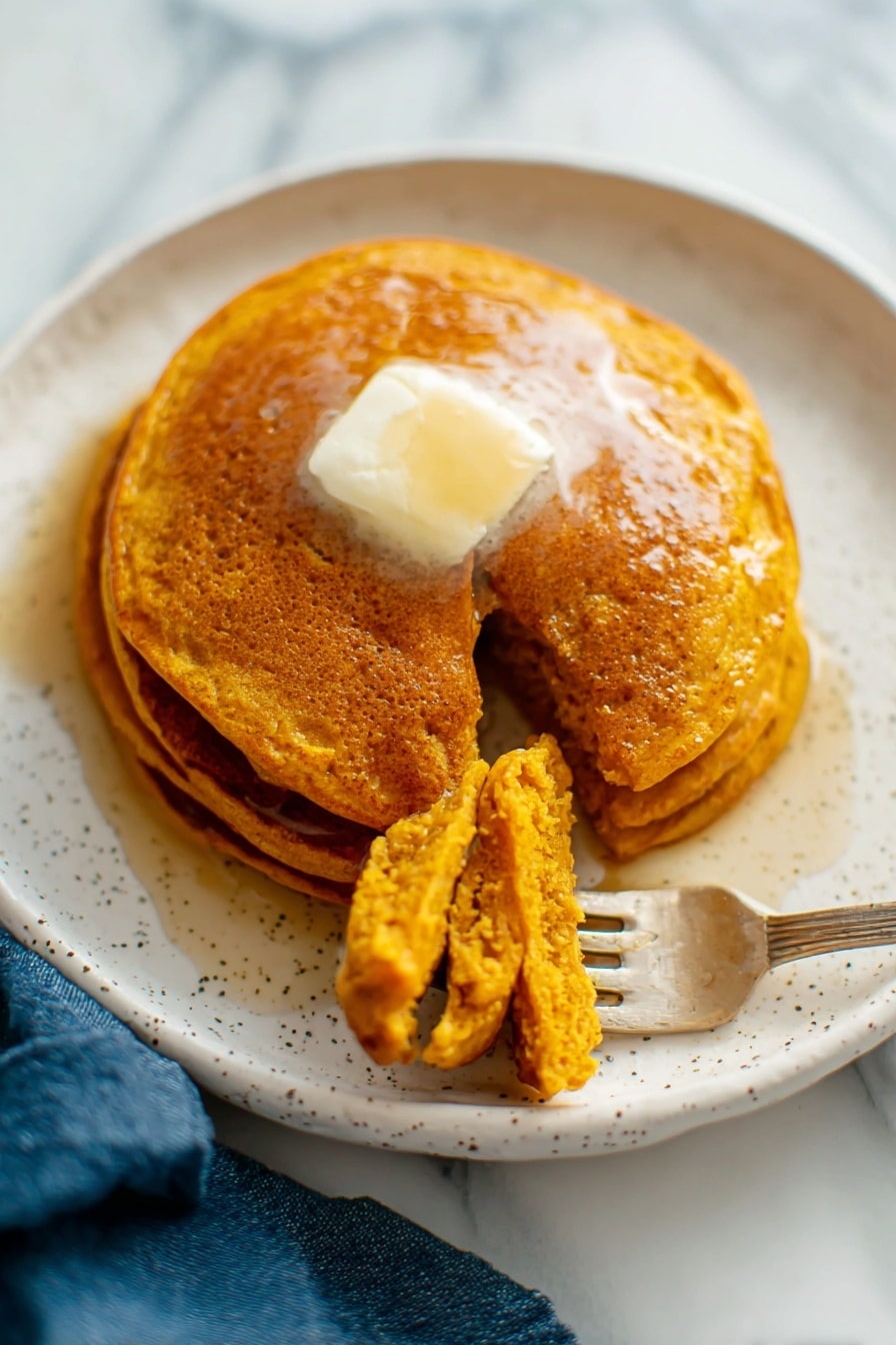 There is a white plate with three golden-brown pancakes stacked slightly unevenly with a clear textured top pancake showing tiny holes. A gold fork rests on the top edge of the plate. Next to the plate on the white marbled surface, there is a pile of more pancakes on another white plate that shows their soft, porous texture. A small white jug with dark syrup is near the plates, and a blue cloth is placed on the surface beside the plate. The photo taken with an iphone --ar 2:3 --v 7 - Whole Wheat Pumpkin Pancakes, healthy pumpkin pancake recipe, fluffy pumpkin pancakes, autumn breakfast ideas, wholesome fall breakfast