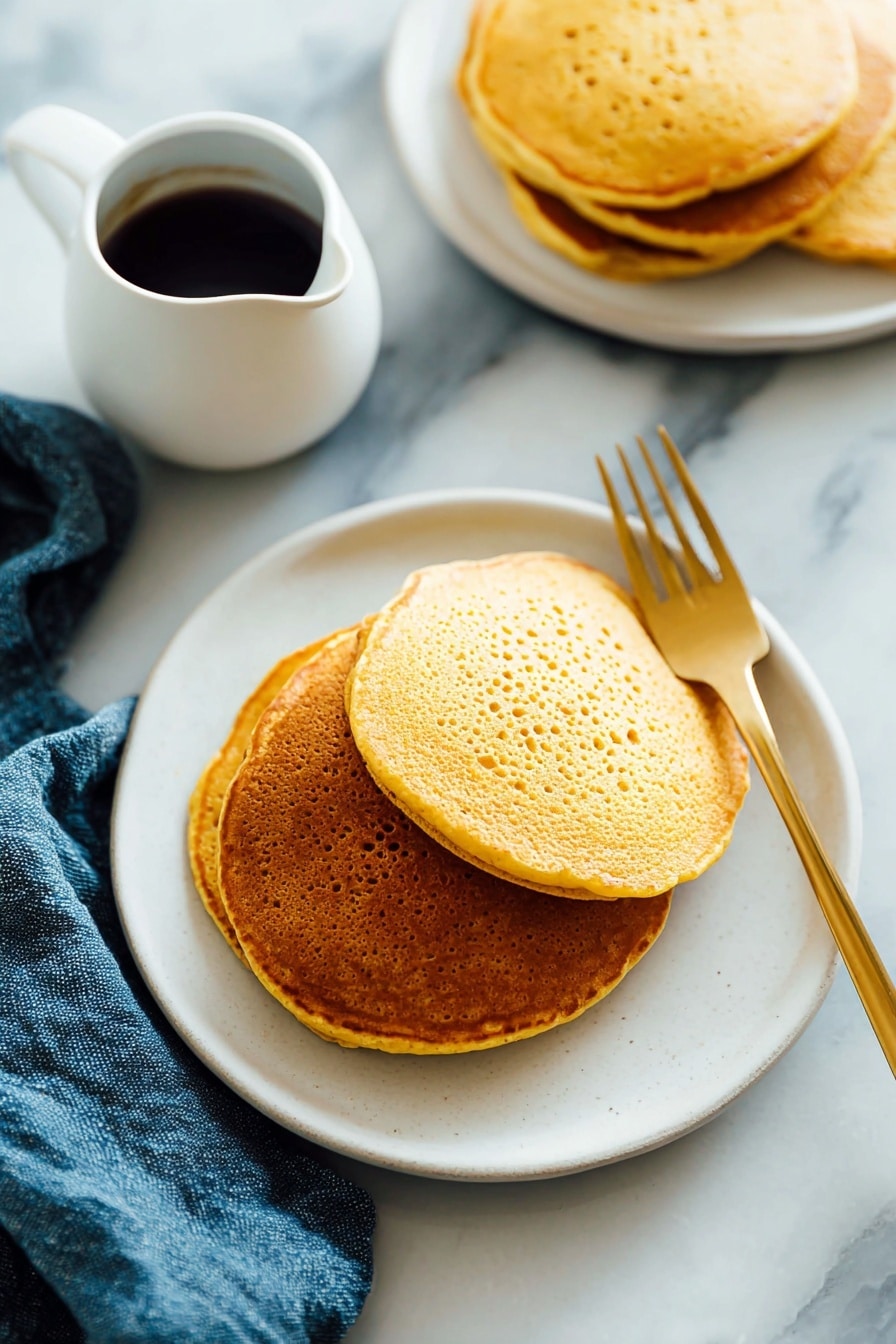 There is a stack of three golden-brown pancakes on a white plate. The pancakes have a slightly porous texture and are arranged with the largest at the bottom and the smallest on top. A light amber syrup is poured over the top pancake, dripping down the sides and pooling a little on the plate. A silver fork rests under the stack, on the left side, and the plate sits on a white marbled surface with a blue cloth nearby. The lighting is soft and natural, highlighting the warm tones of the pancakes and syrup. photo taken with an iphone --ar 2:3 --v 7 - Whole Wheat Pumpkin Pancakes, healthy pumpkin pancake recipe, fluffy pumpkin pancakes, autumn breakfast ideas, wholesome fall breakfast