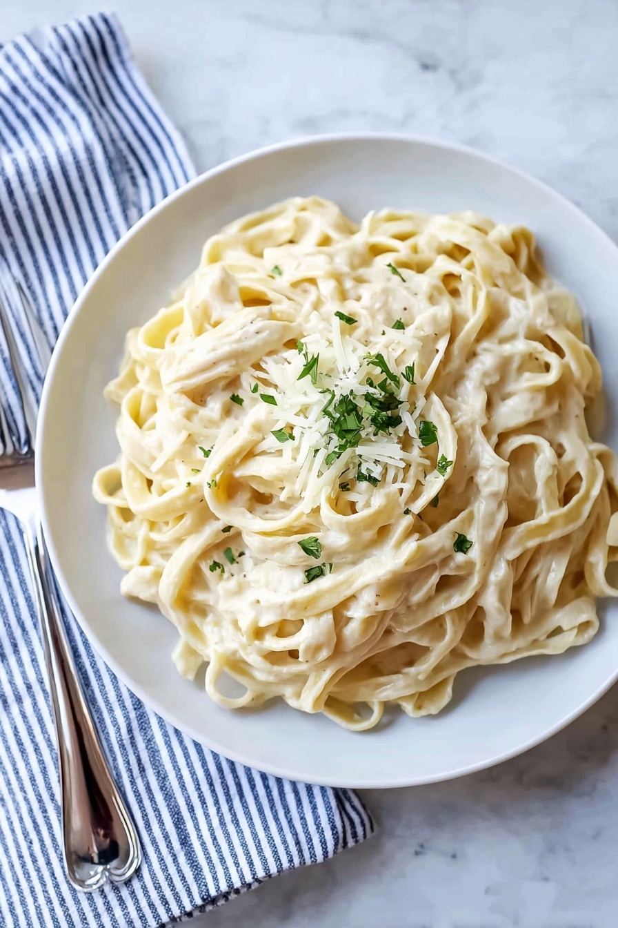 A white plate filled with a large serving of creamy fettuccine pasta, with smooth, thick noodles coated in a pale, off-white sauce. On top in the center, there is a small pile of grated white cheese and finely chopped green herbs adding a touch of color. The dish sits on a white marbled surface, with a silver fork resting on a striped blue and white cloth to the left side of the plate. The lighting is soft and natural, highlighting the creamy texture of the pasta and the freshness of the garnish. photo taken with an iphone --ar 2:3 --v 7 - Creamy Fettuccine Alfredo, easy creamy pasta, Italian pasta recipe, comforting dinner idea, classic Alfredo sauce