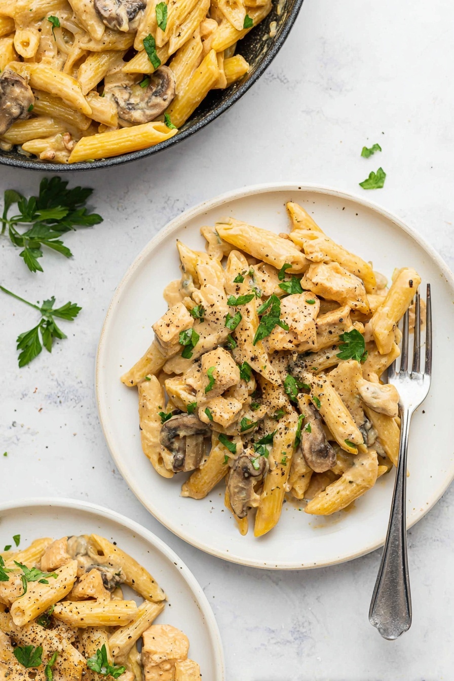 A white plate holds creamy pasta made of penne noodles mixed with pieces of cooked chicken and sliced mushrooms. The pasta and chicken are coated with a light beige creamy sauce, giving a smooth texture. The mushrooms are brown with soft white inside visible, placed evenly throughout. Green parsley leaves are sprinkled on top, adding a fresh color contrast, and there are small bits of red pepper flakes and black pepper scattered over the dish. A silver fork is partly stuck into the pasta on the right side, resting on the plate. The whole scene is set on a white marbled surface, creating a clean and bright background. photo taken with an iphone --ar 2:3 --v 7 - Creamy Chicken Marsala Pasta, Chicken Marsala pasta, creamy chicken pasta, Italian chicken pasta recipe, quick chicken pasta dish