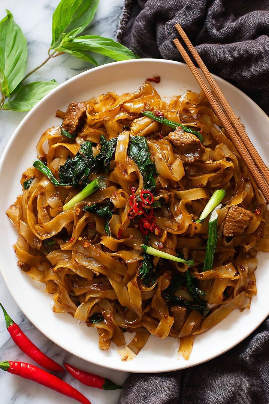A white plate filled with flat brown noodles mixed with glossy pieces of cooked meat that have a caramel color. Scattered through the noodles are bright green pieces of vegetables, including spring onions with white and green parts, and dark leafy greens, all coated in a slightly oily sauce with hints of red chili flakes. A pair of wooden chopsticks rests on the edge of the plate, gripping some noodles and meat. The plate sits on a white marbled surface with a dark grey cloth draped underneath and small bright red chili peppers with fresh green leaves nearby. Photo taken with an iphone --ar 2:3 --v 7 - Easy Spicy Drunken Noodles with Chicken, Thai drunken noodles, spicy chicken stir-fry, quick Thai noodle recipe, flavorful chicken noodle dish