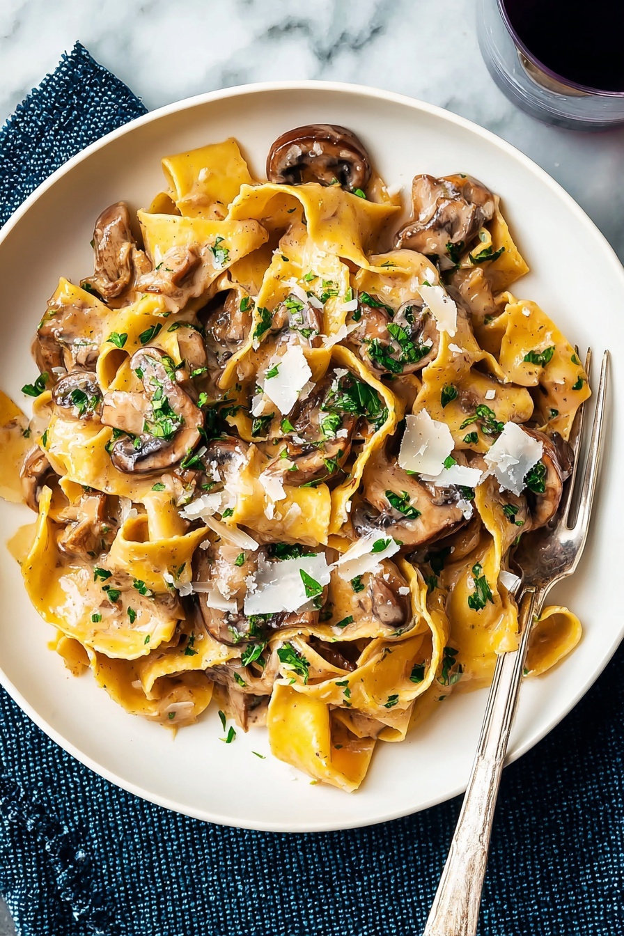 A white plate holds a serving of wide, yellow pasta ribbons cooked in a light brown creamy mushroom sauce. On top, there are several brown, slightly browned mushroom slices mixed throughout the pasta, adding a smooth texture. Bright green chopped herbs are sprinkled over the dish, giving a fresh contrast. Light shavings of white cheese are scattered on top, melting slightly into the sauce. A silver fork rests on the edge of the plate, and the plate is placed on a white marbled surface with a dark blue textured cloth beneath one side. Photo taken with an iphone --ar 2:3 --v 7 - Vegetarian Mushroom Stroganoff, mushroom stroganoff recipe, easy vegetarian dinner, creamy mushroom sauce, quick weeknight vegetarian meal