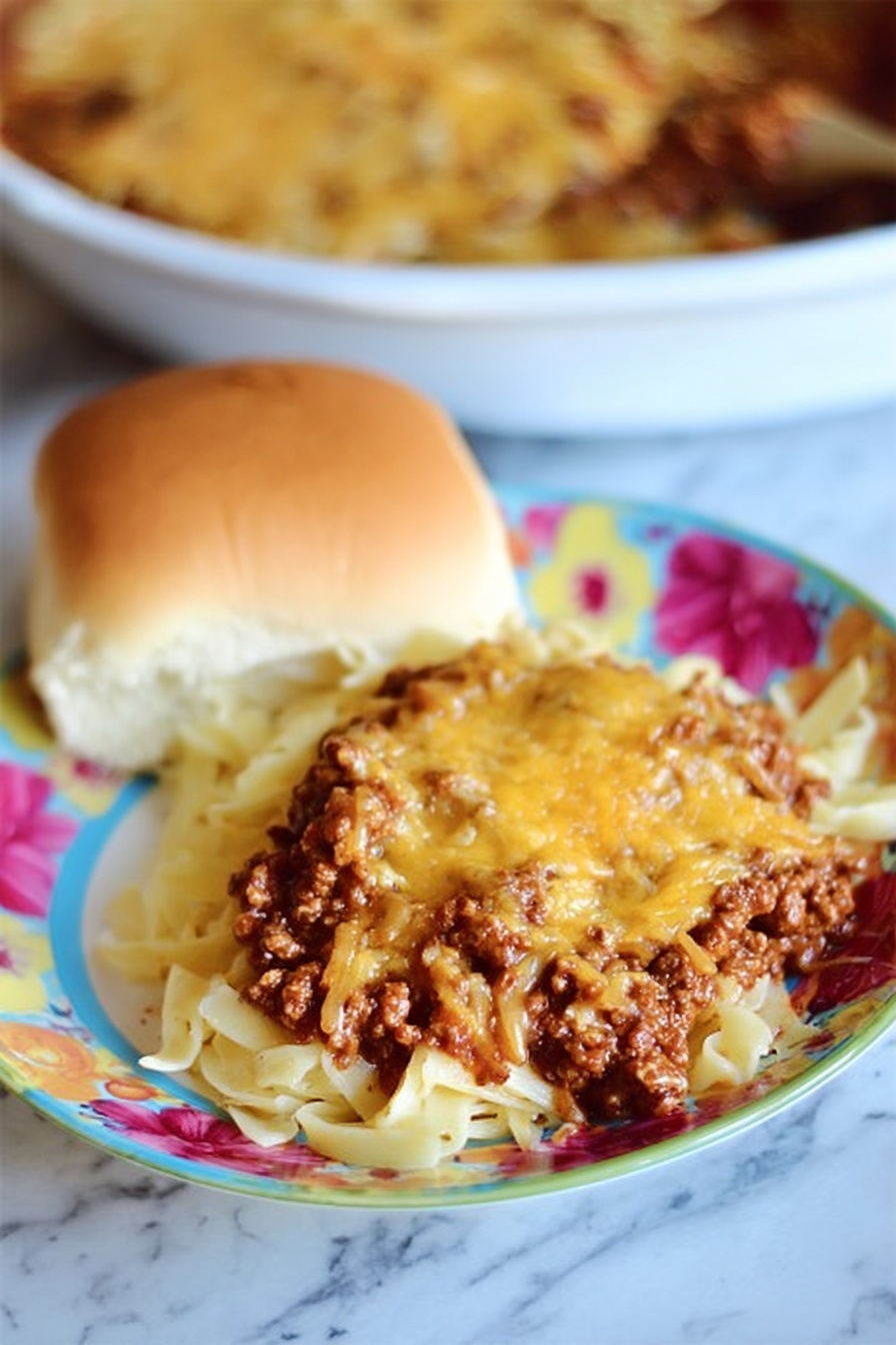 The dish shows a serving of pasta on a white plate with a colorful flower design in pink, red, and yellow. The pasta forms the bottom layer with visible white and slightly curved noodles. On top of the pasta, there is a thick layer of cooked ground meat in dark brown color mixed with reddish-brown sauce. The top layer is melted orange-brown cheese covering most of the meat. Next to the pasta is a soft, golden-brown bread roll with a smooth surface and light cracks. The background shows a white round dish filled with the same pasta, meat, and cheese mix. The whole scene is set on a white marbled surface. Photo taken with an iphone --ar 2:3 --v 7 - Sour Cream Noodle Bake, Easy Ground Beef Casserole, Creamy Baked Noodles, Cozy Weeknight Dinner, Cheesy Pasta Bake