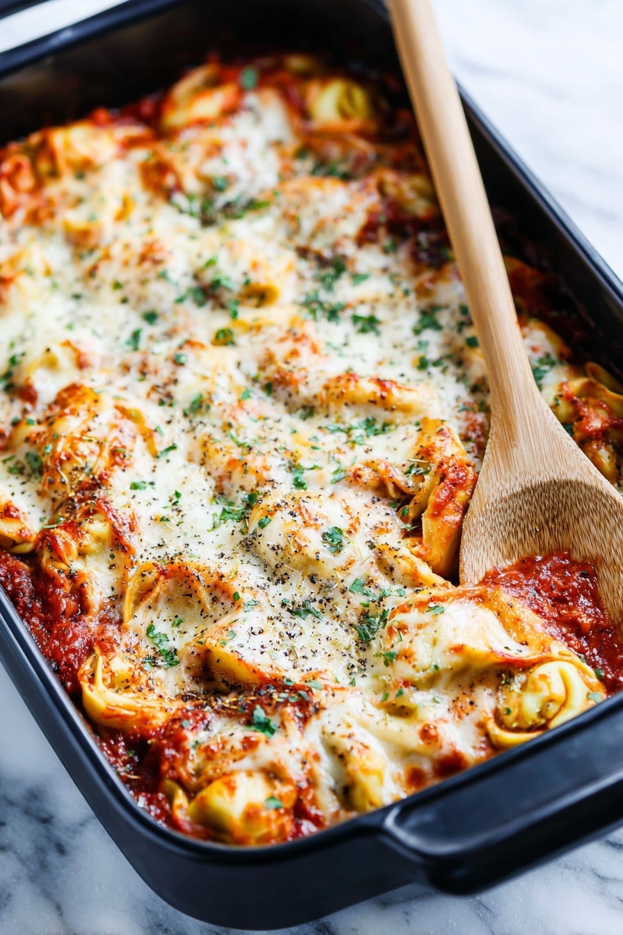 A black baking dish holds a layered tortellini pasta bake with a rich tomato sauce. The top layer is melted white cheese, bubbly and slightly browned in spots, mixed with twisted yellow tortellini filled pasta visible underneath. The sauce is deep red with bits of herbs and small chunks of meat mixed in. Scattered green herb leaves add color contrast across the top. The dish rests on a white marbled surface with a folded striped cloth nearby and a wooden-handled black spoon on the right edge of the frame, creating a cozy kitchen feel. photo taken with an iphone --ar 2:3 --v 7 - Cheesy Baked Tortellini Casserole, baked tortellini recipes, cheesy pasta casserole, hearty tortellini dinner, easy baked pasta dishes