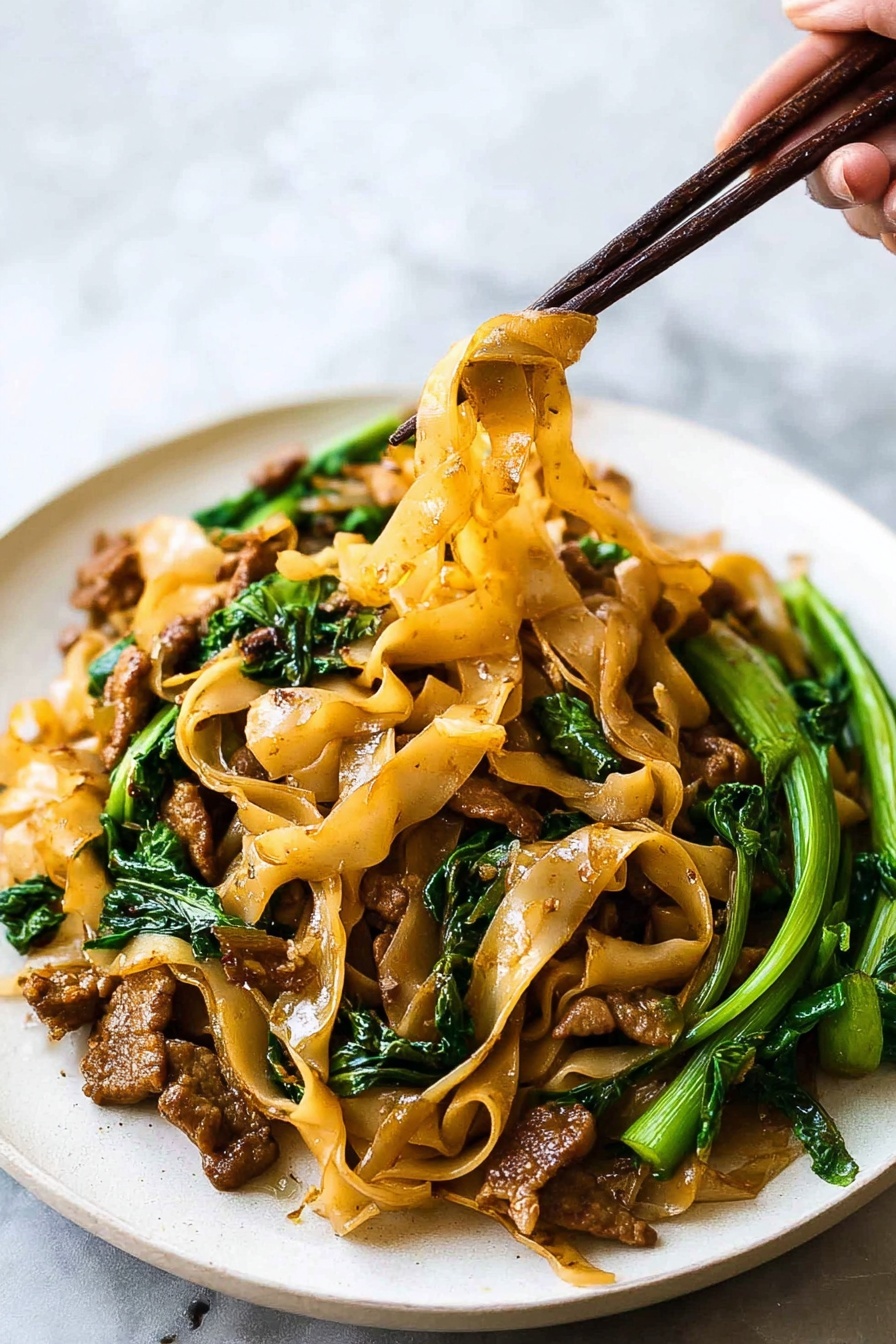 This image shows a white plate filled with wide flat light brown noodles mixed with small pieces of browned meat and bright green leafy vegetables. The noodles look soft and slightly oily, hanging in strands from dark brown chopsticks held by a woman's hand. The meat pieces are scattered throughout the dish, with some near the edges and some under the noodles. The green vegetables add a fresh look, with stems and leaves placed mostly on the right side of the plate. The background is a soft white marbled texture, keeping the focus on the colorful noodles and greens. Photo taken with an iphone --ar 2:3 --v 7 - Thai Pad See Ew, easy Pad See Ew recipe, authentic Thai noodles, quick Thai stir-fry, homemade Thai street food