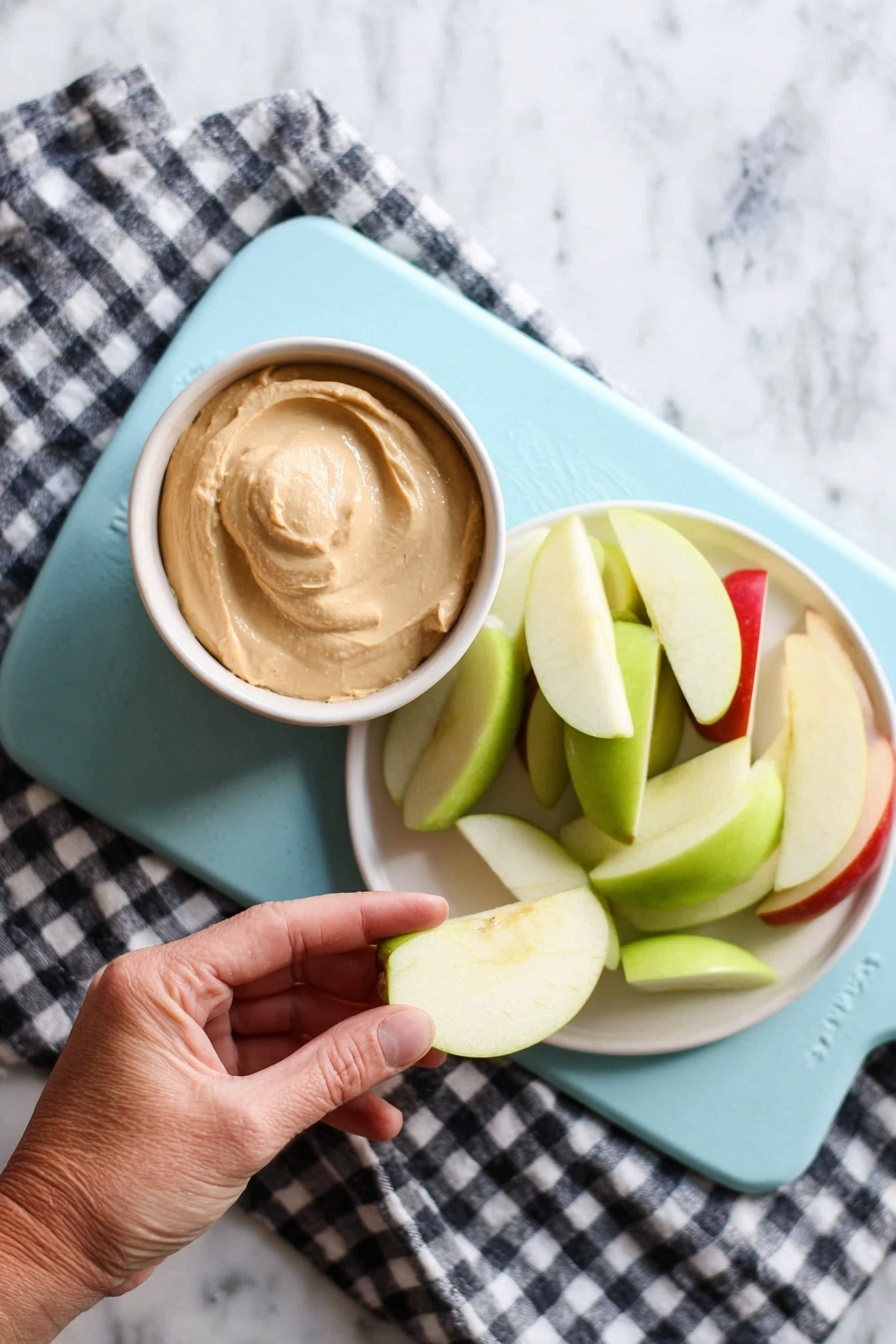 A woman's hand is holding a pale green apple slice dipped in a small bowl of light brown creamy dip. Next to it, on a white plate, there is a pile of apple slices in green and red colors. Both the bowl and plate are placed on a light blue cutting board, which rests on a black and white checkered cloth on a white marbled surface. The scene is brightly lit, showing clear textures of the creamy dip and crisp apple slices. photo taken with an iphone --ar 2:3 --v 7 - Caramel Apple Dip, caramel apple dip, creamy caramel dip, apple dip recipe, easy caramel dip