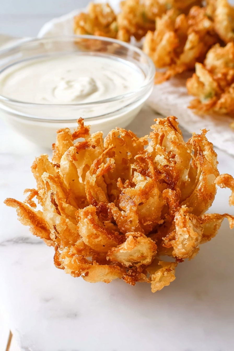A close-up image of a golden-brown, crispy fried onion blossom placed on a white marbled surface. The onion blossom is fully opened like a flower with many uneven, crunchy petals showing a textured, fried batter coating. Next to it is a small clear glass bowl filled with smooth, creamy white dipping sauce. In the background, some more fried onion blossoms are slightly blurred. The scene is bright and simple with clean white tones and a soft shadow under the food. photo taken with an iphone --ar 2:3 --v 7 - Homemade Blooming Onion, crispy blooming onion recipe, easy onion appetizer, homemade onion petals, spicy dipping sauce