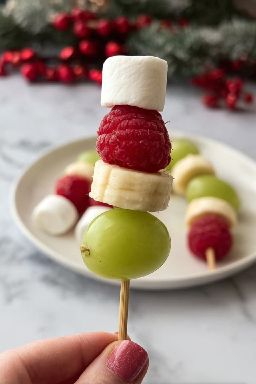 A small skewer held by a woman's hand shows four layers: at the bottom, a smooth, green grape; above it, a round, pale yellow banana slice; next a bright red raspberry with visible bumpy texture; and on top, a small white marshmallow shaped like a cube. In the background, more similar skewers rest on a white plate on a white marbled surface with some red berries. The overall look is fresh, colorful, and festive. photo taken with an iphone --ar 2:3 --v 7 - Grinch Fruit Kabobs, holiday fruit skewers, festive snack ideas, kid-friendly party treats, colorful fruit kabobs