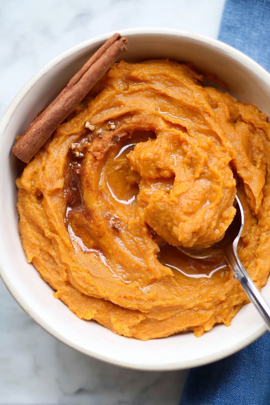 The image shows a white bowl filled with thick, smooth orange sweet potato mash. The mash has swirled texture with a shiny surface, topped with a light drizzle of syrup pooling slightly in the center. A cinnamon stick rests on the left side of the mash, and small bits of brown spices are sprinkled on top. A metal spoon is scooping up part of the mash on the right side, with the spoon handle extending out of the bowl. The bowl sits on a white marbled surface with a bit of a blue cloth visible in the bottom right corner. Photo taken with an iphone --ar 2:3 --v 7 - Brown Butter Mashed Sweet Potatoes, mashed sweet potatoes with brown butter, spiced sweet potato side dish, holiday sweet potato recipes, easy sweet potato sides